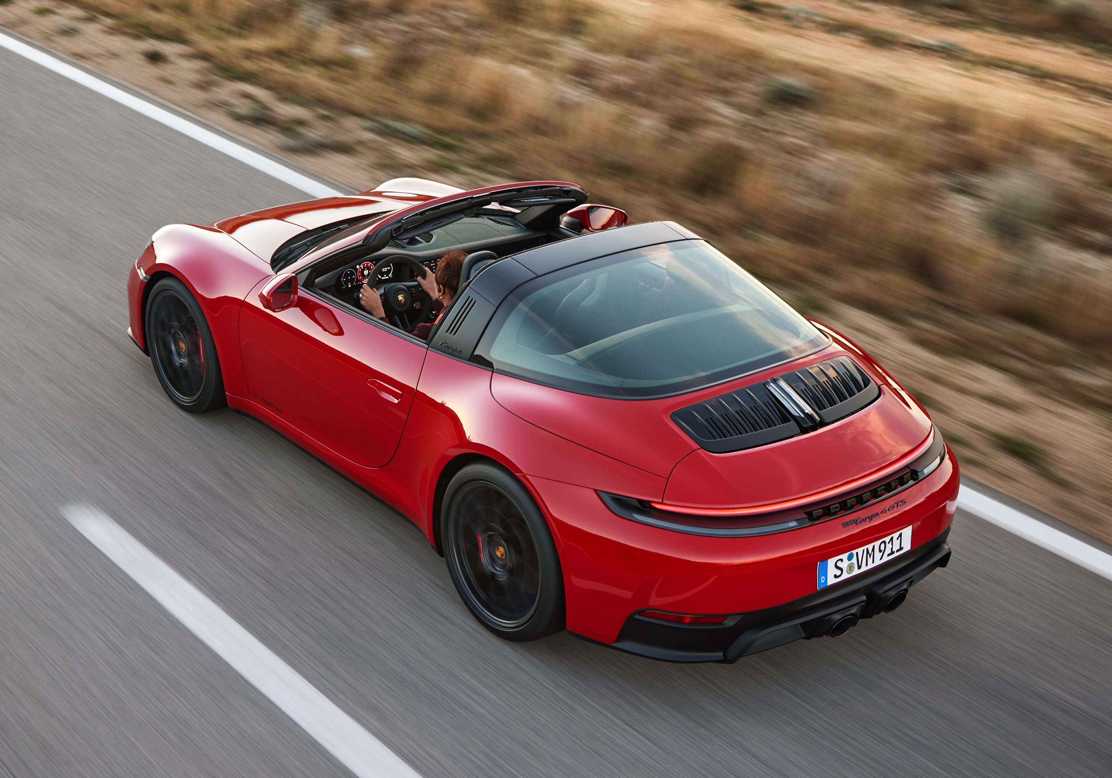 Красный спортивный автомобиль Porsche 911 Targa с черным складным верхом, в движении по асфальтированной дороге с белой разметкой, по бокам - сухая растительность, размытая движением.