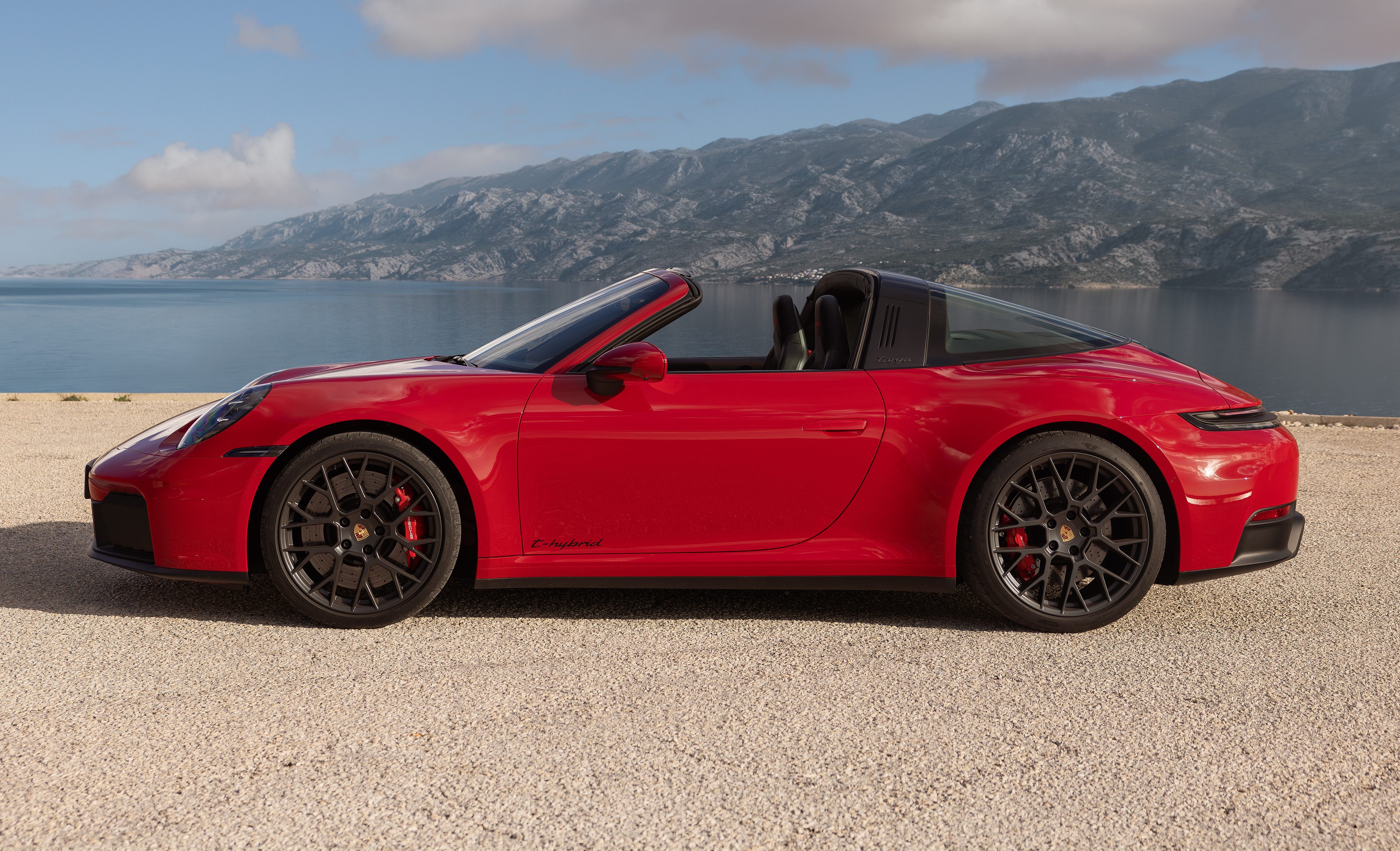 Carro esportivo vermelho, um Porsche 911 Targa, estacionado em uma estrada de terra com o teto retrátil aberto. Ao fundo, um lago azul e montanhas sob um céu claro com nuvens brancas