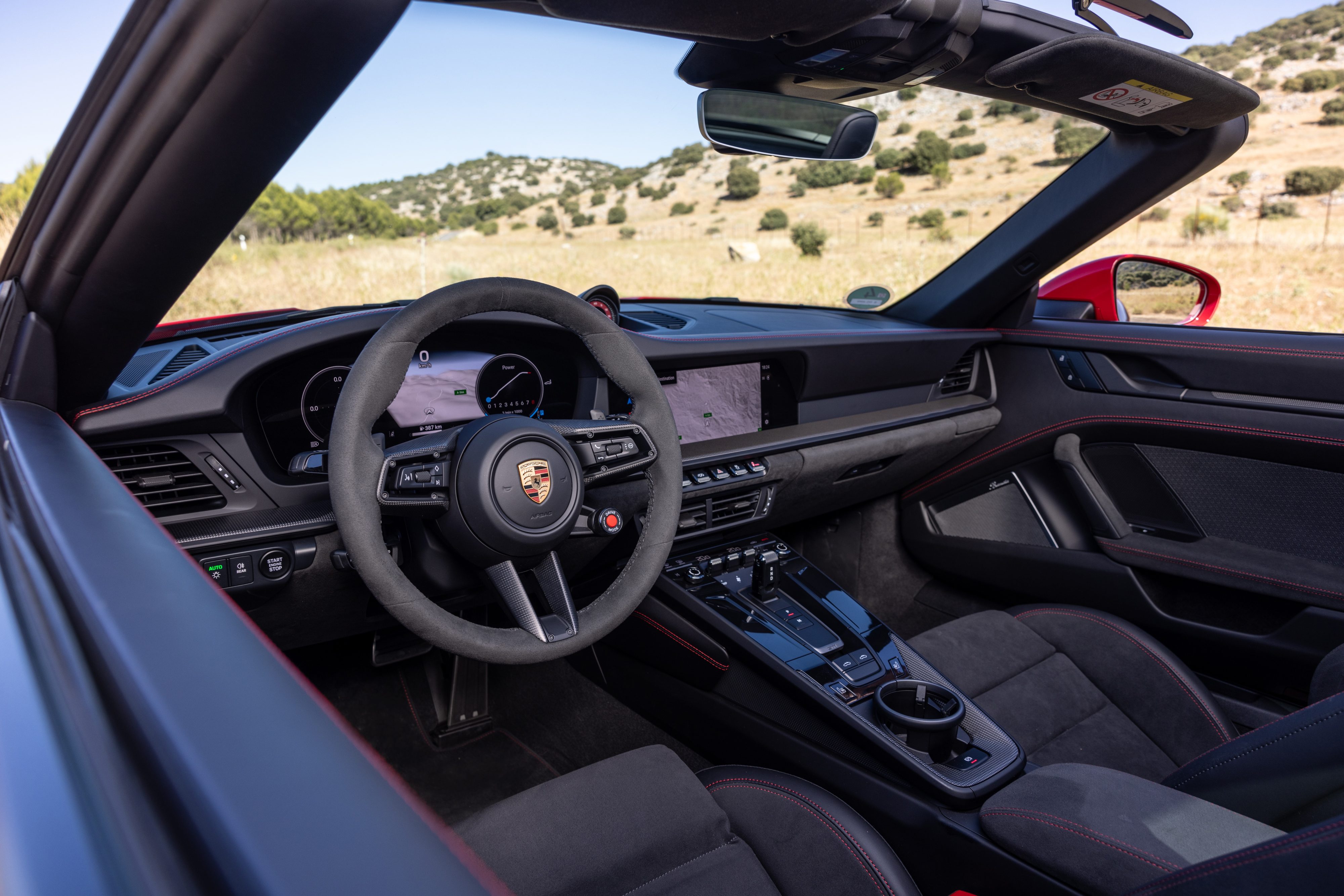 Interior de um carro esportivo conversível vermelho, com o volante e painel escuros em primeiro plano. O volante tem o emblema da Porsche. Ao fundo, uma paisagem montanhosa árida sob céu azul claro