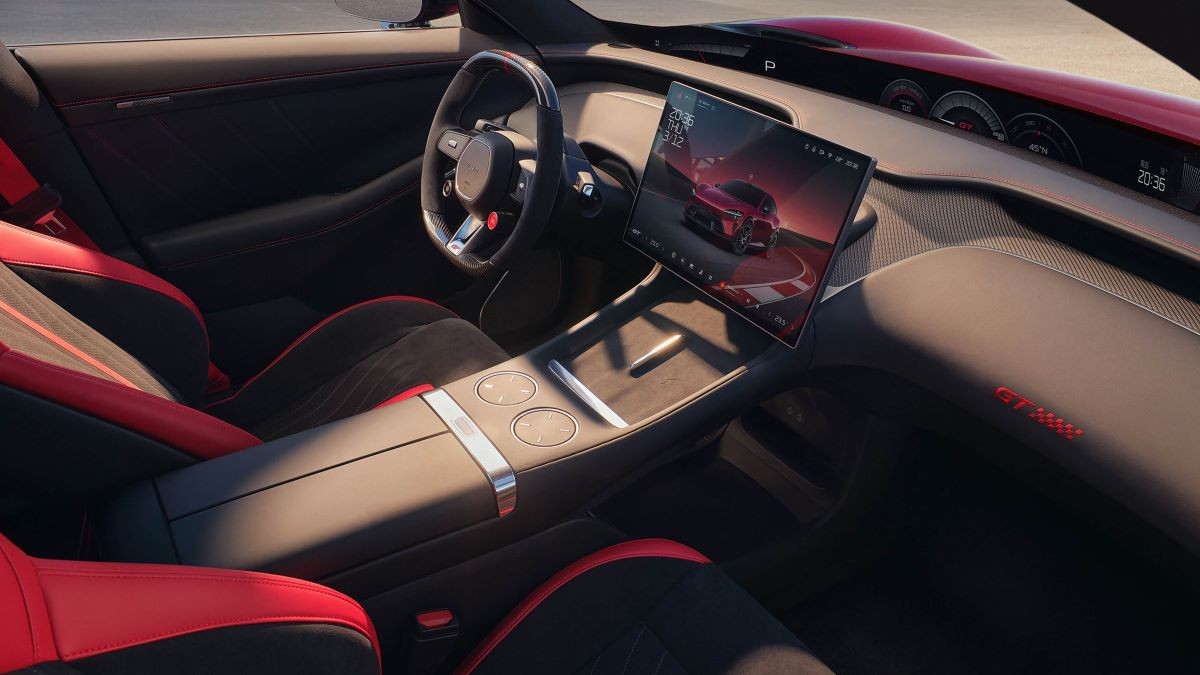 Interior de um carro esportivo moderno com bancos pretos e detalhes vermelhos, volante esportivo, console central com porta-copos e uma tela multimídia grande exibindo um carro vermelho em uma pista de corrida