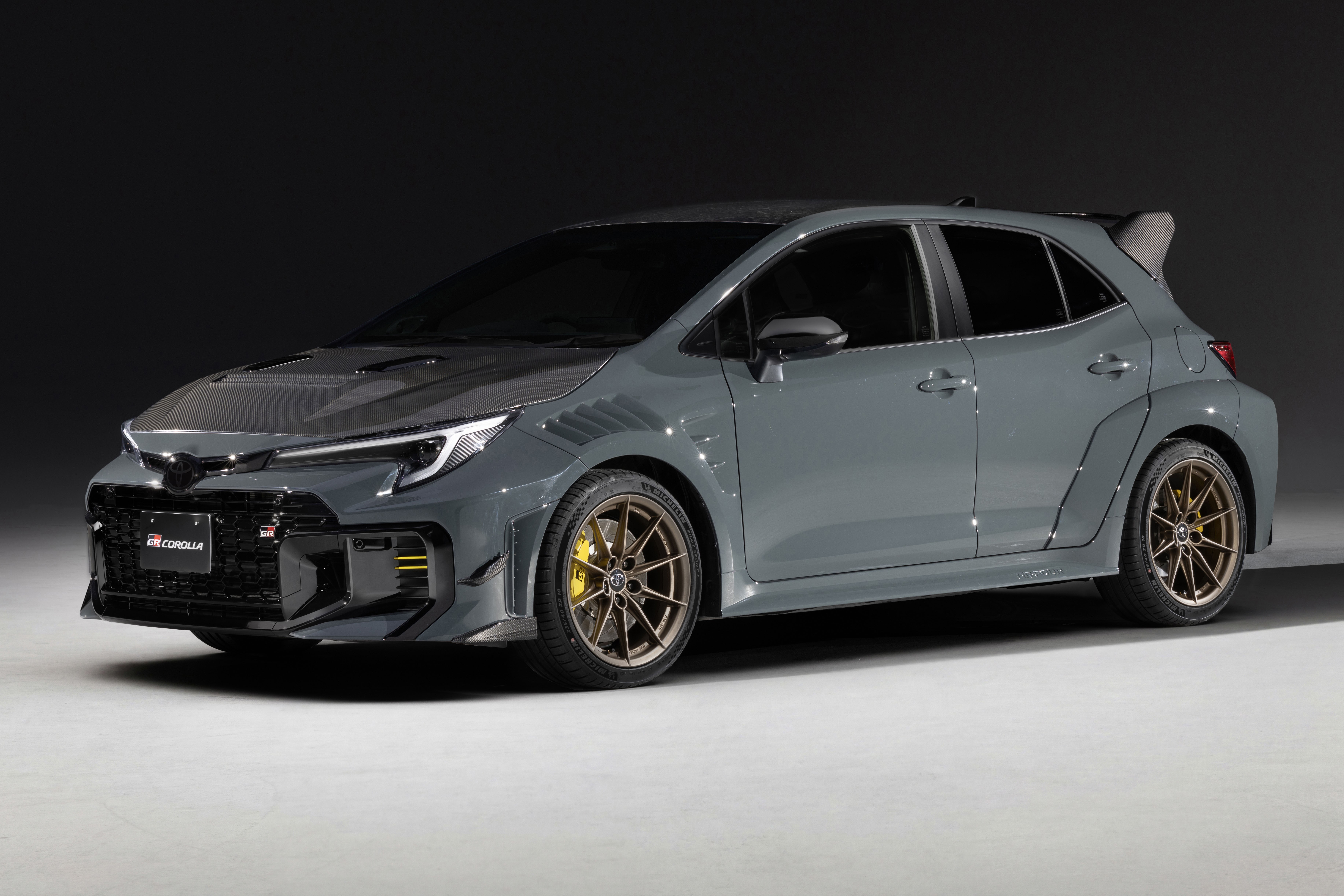 Carro esportivo cinza-chumbo, modelo hatchback, com capô escuro e rodas douradas, parado em estúdio com fundo escuro
