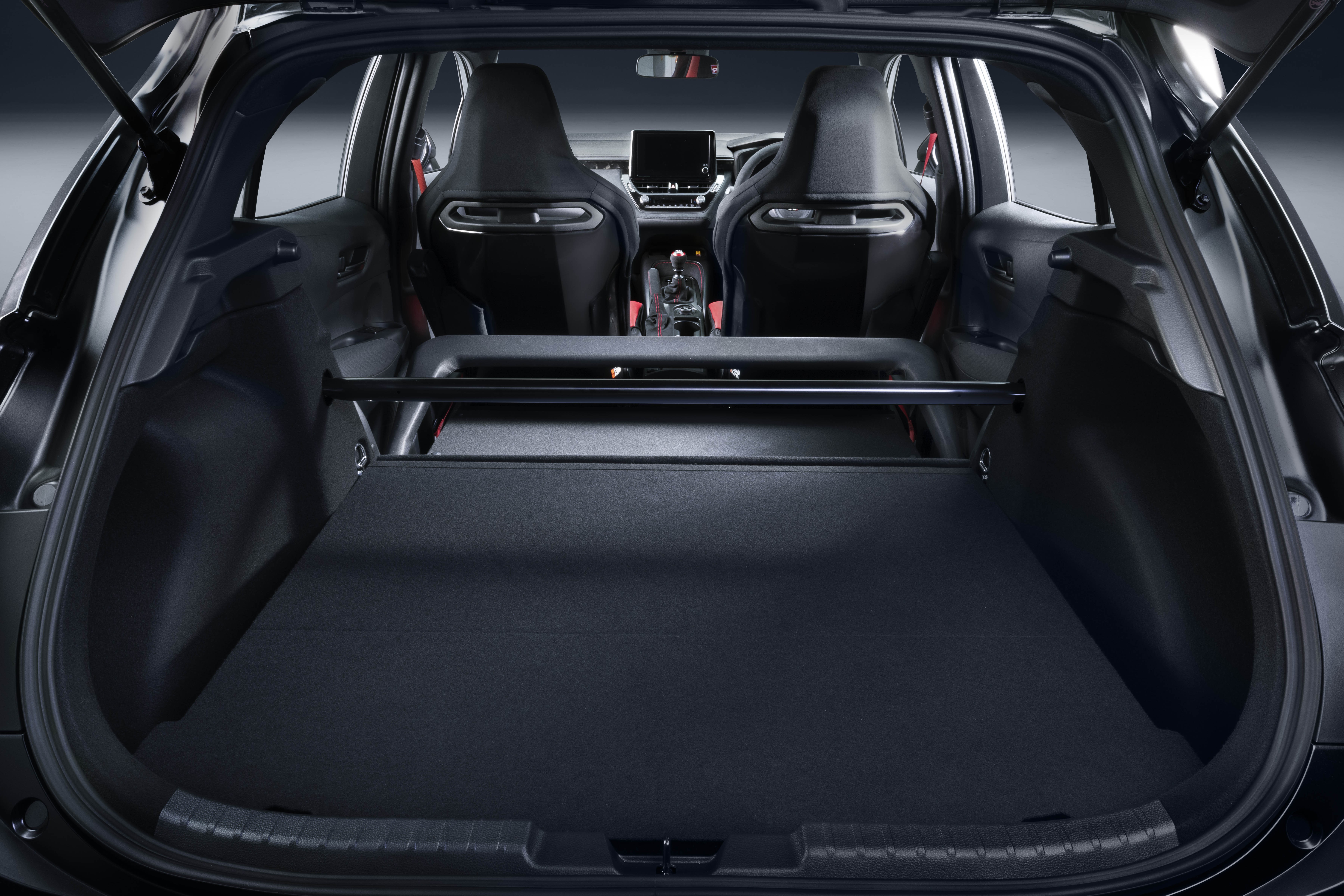Interior de um carro esportivo preto, visto do porta-malas aberto. O porta-malas tem um assoalho plano e uma barra de reforço preta. À frente, dois bancos esportivos pretos com detalhes prateados e o painel do carro com uma tela central. O ambiente é escuro e focado nos detalhes internos do veículo