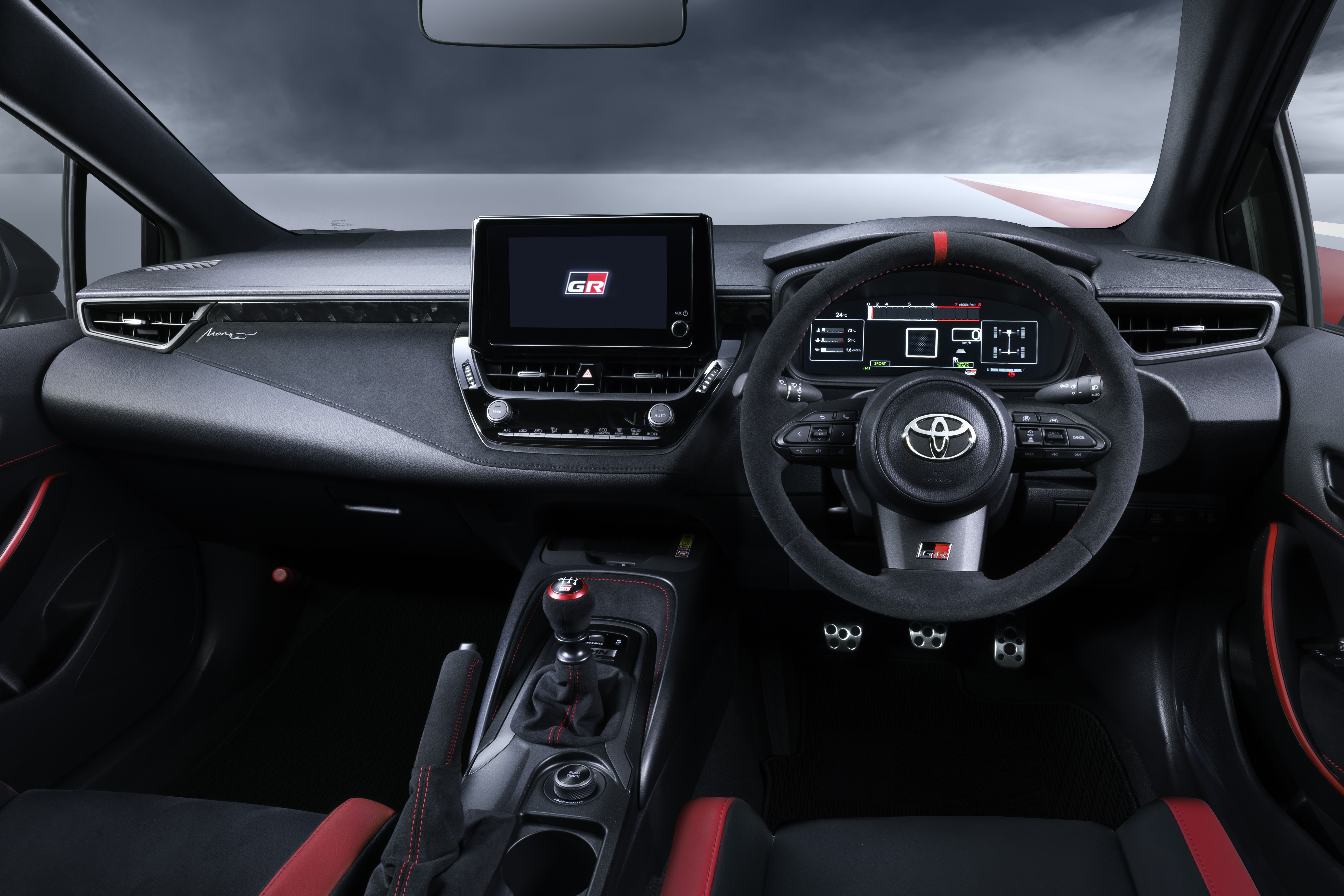 Interior de um carro esportivo com volante de camurça preta e faixa vermelha, painel digital, tela multimídia com logo GR, câmbio manual e bancos pretos com detalhes vermelhos