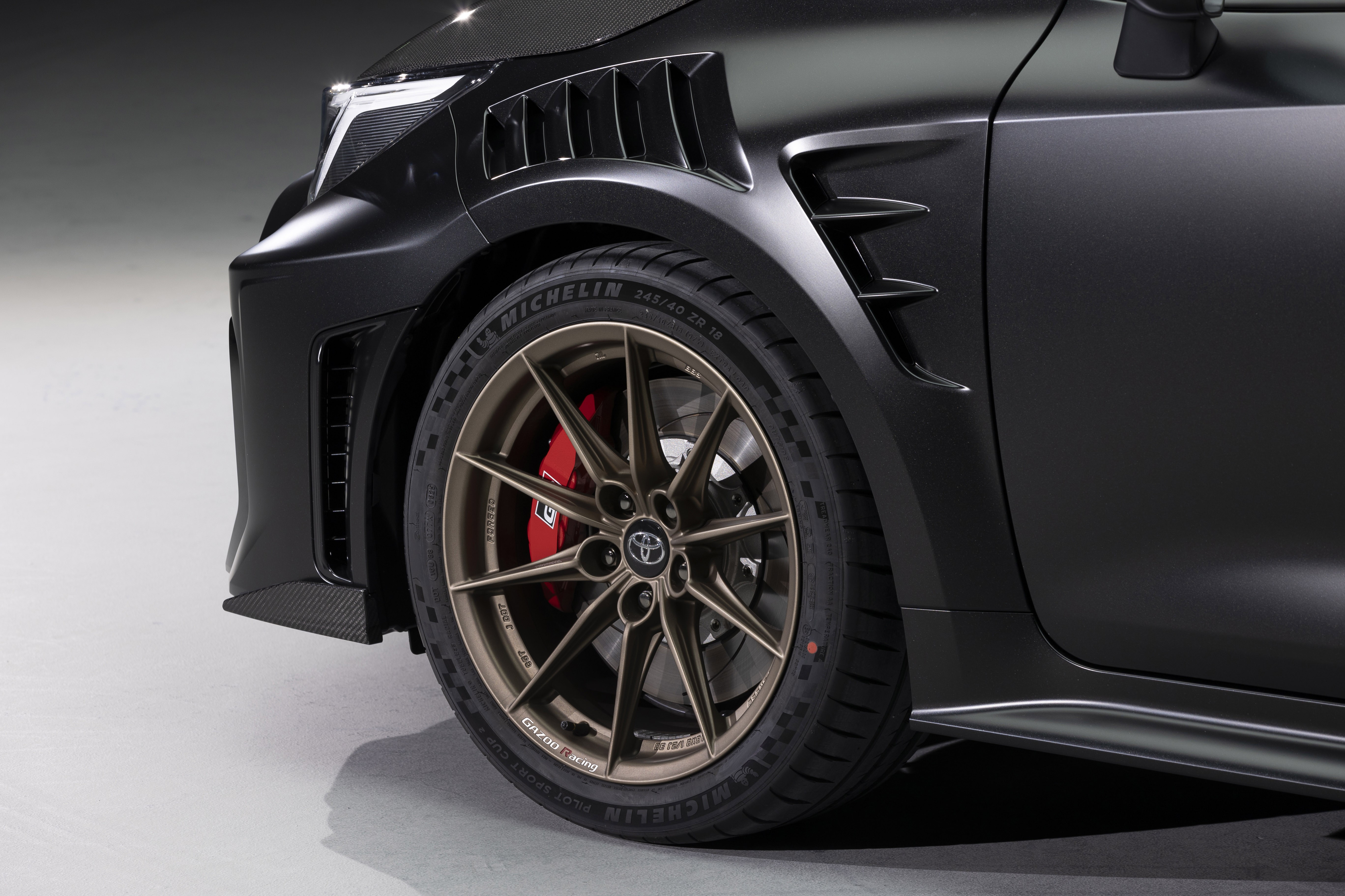 Lateral dianteira de um carro esportivo preto fosco, com farol LED branco aceso, grade frontal e saídas de ar no para-lama. A roda de liga leve bronze tem pinças de freio vermelhas e pneus Michelin