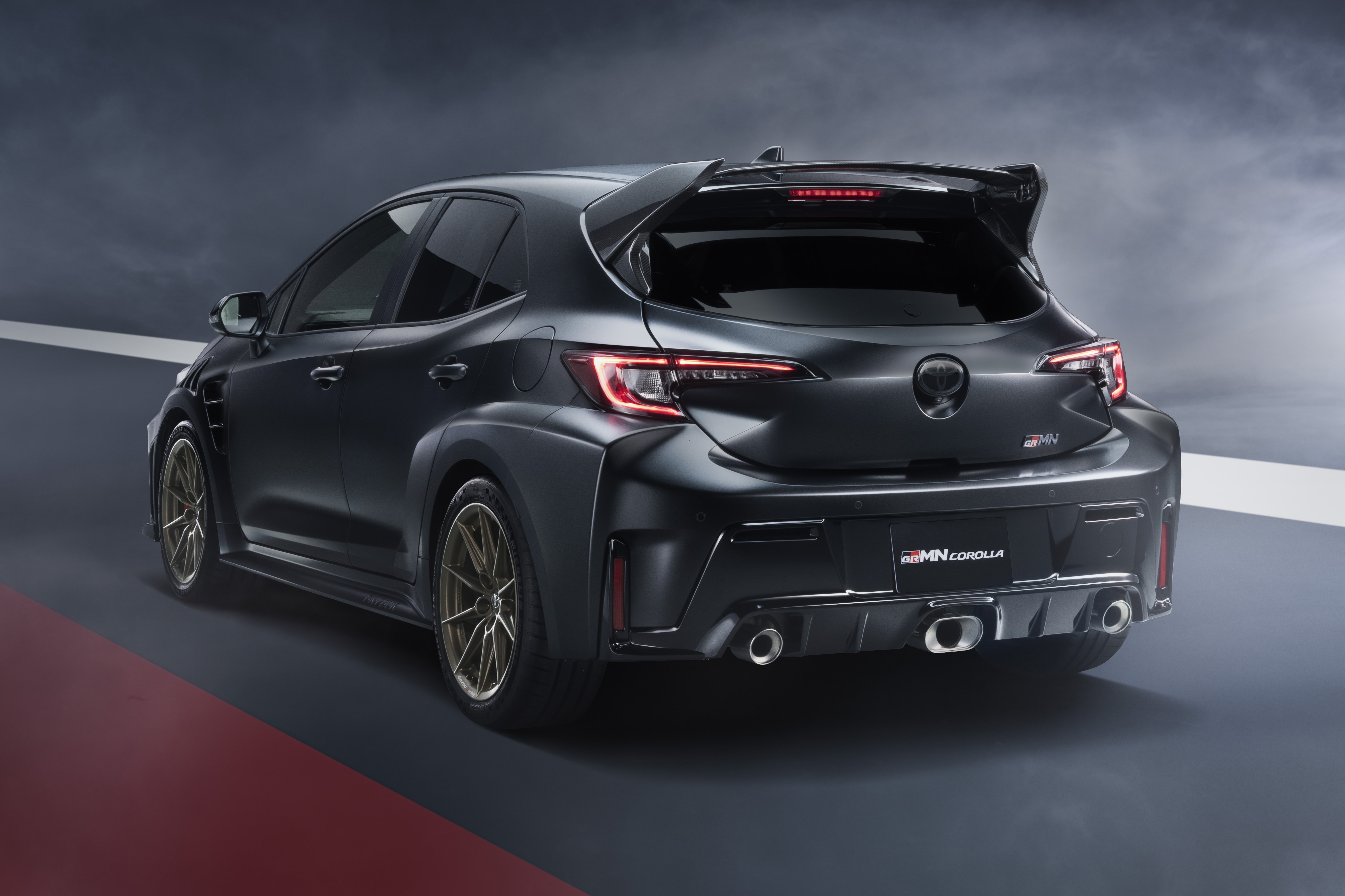 Toyota GRMN Corolla cinza escuro, visto de traseira e lateral, com rodas douradas, aerofólio e três saídas de escapamento, em uma pista escura com faixas vermelha e branca