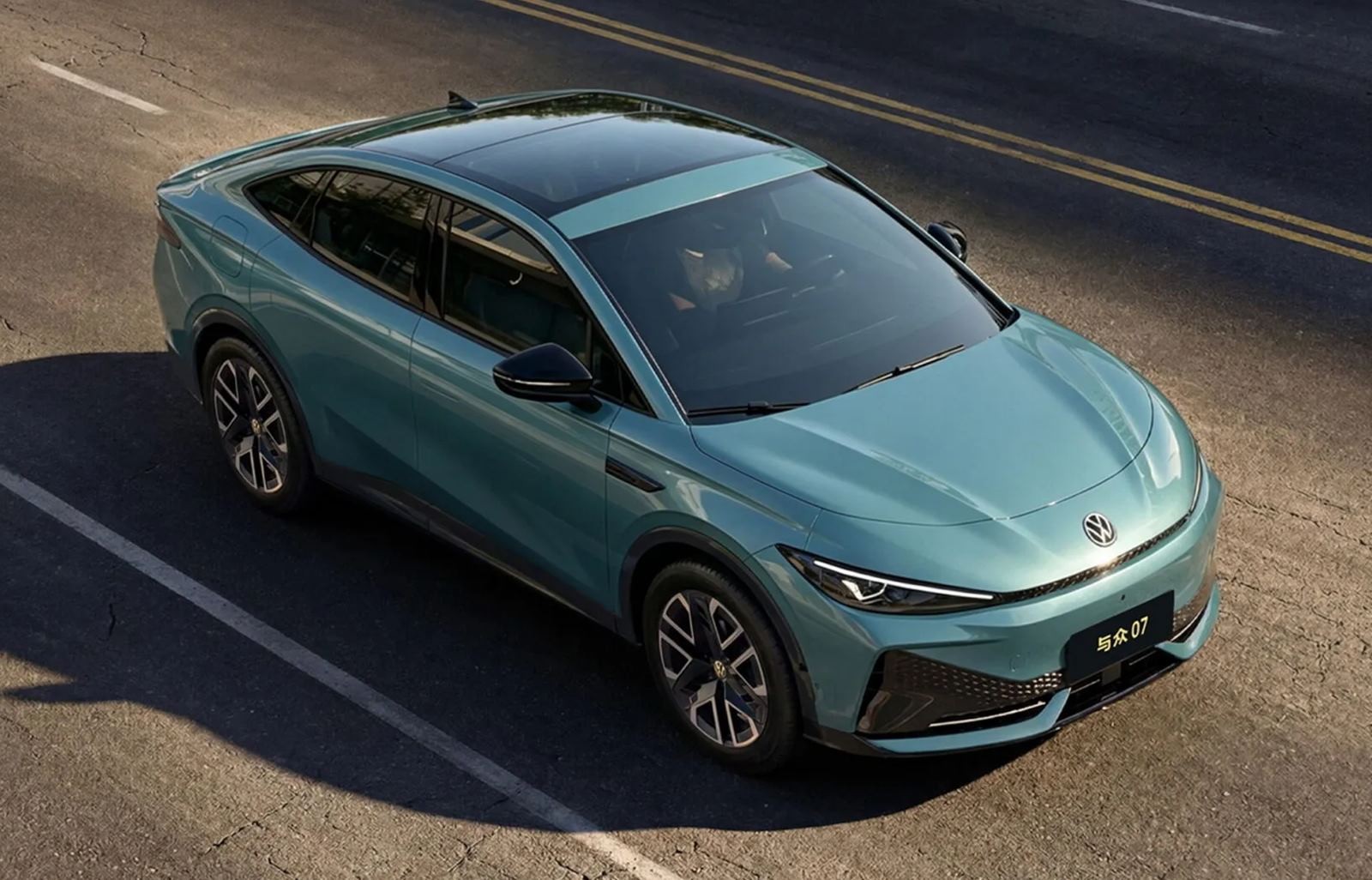 Carro elétrico azul-petróleo, estilo cupê, com teto solar panorâmico, estacionado em uma rua com faixas brancas e amarelas. O motorista é visível através do para-brisa. O veículo possui rodas pretas com detalhes prateados e a placa frontal exibe caracteres chineses