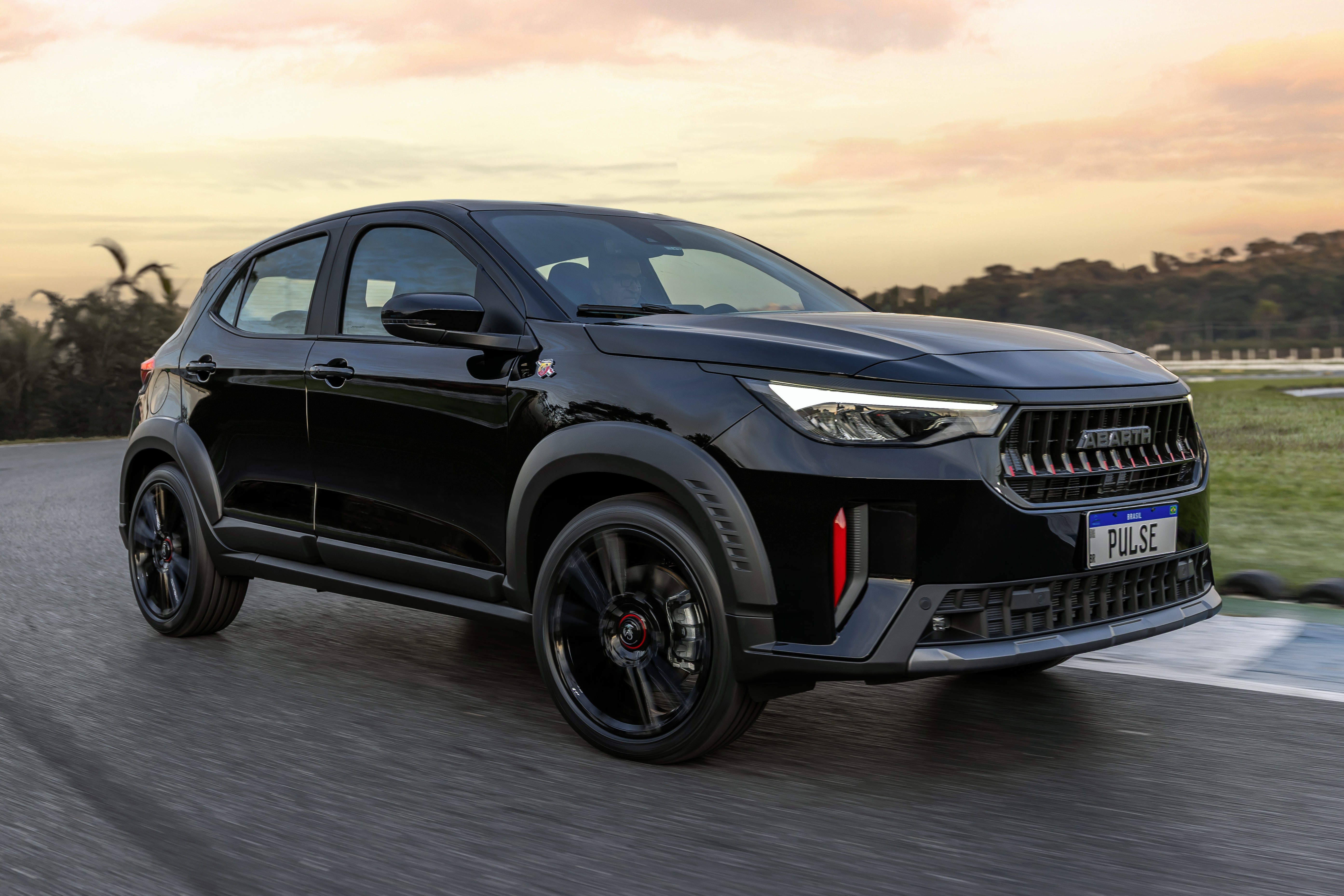 SUV Fiat Pulse Abarth preto brilhante em movimento, visto de perfil frontal, com detalhes vermelhos na grade e para-choque, rodas escuras e placa PULSE azul. O carro está em uma pista de asfalto, com vegetação e céu alaranjado ao fundo