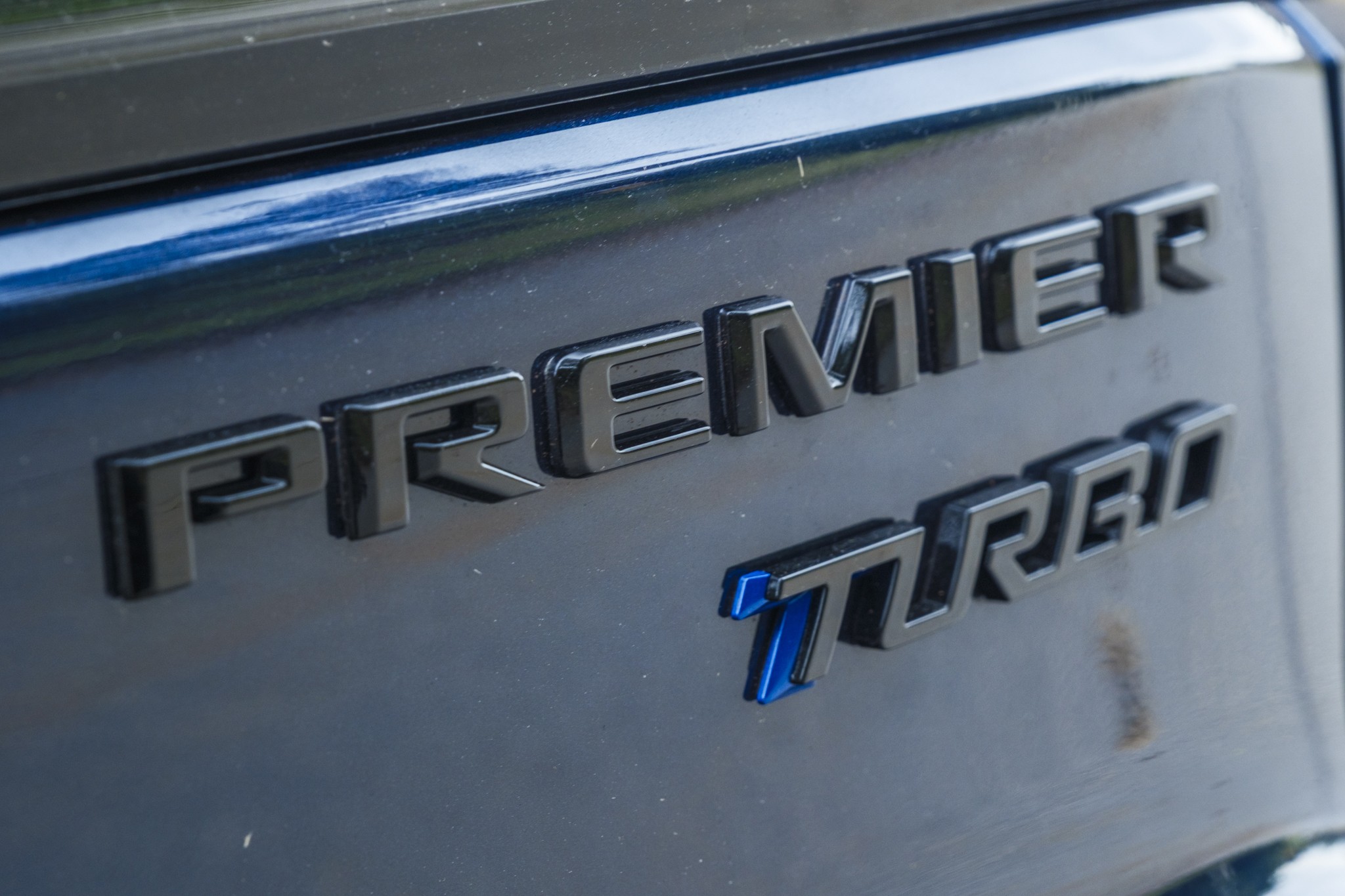 Emblema PREMIER TURBO em relevo preto fosco na traseira de um carro azul escuro, com o T de TURBO em azul vibrante