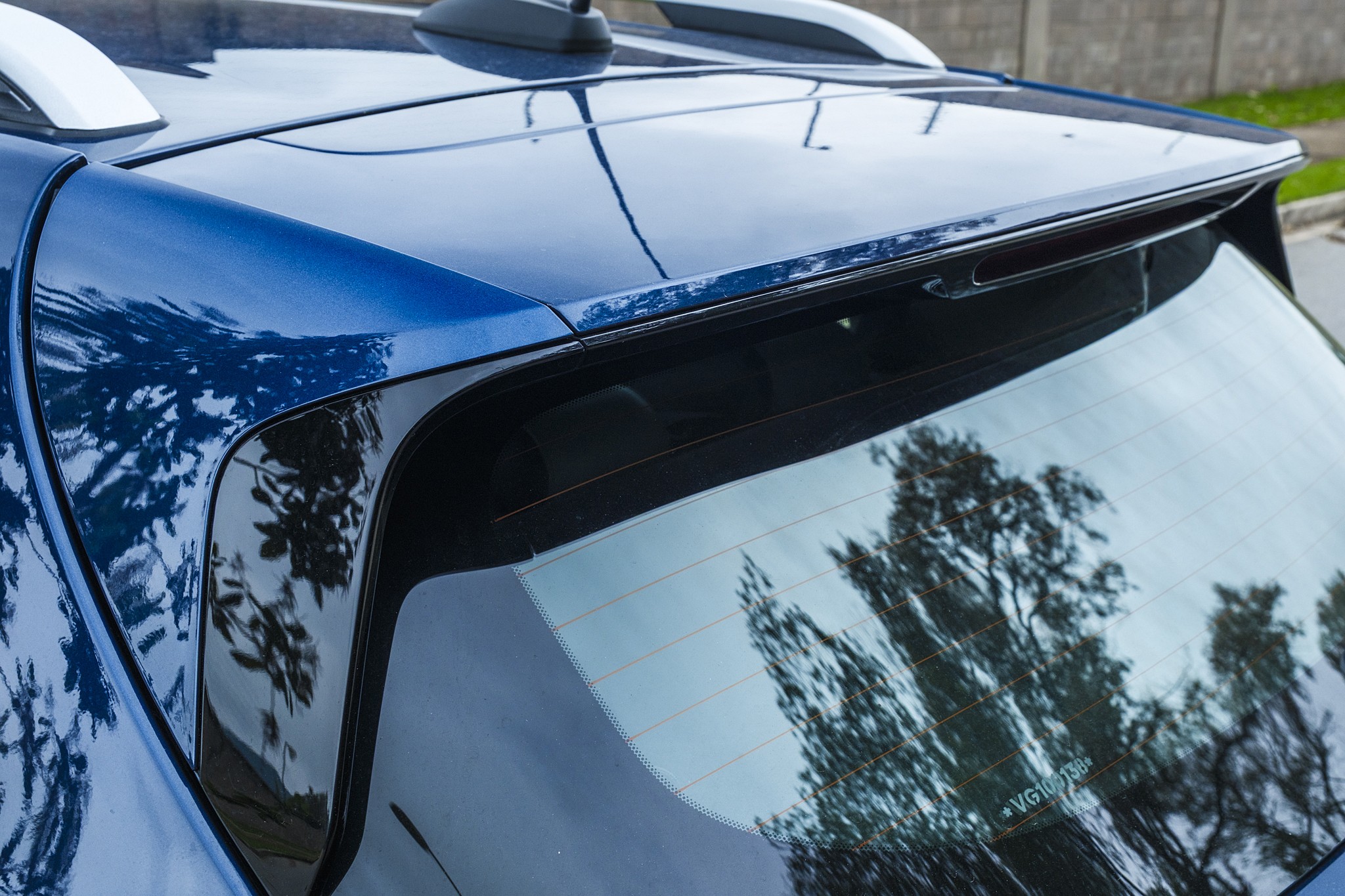 Parte traseira de um carro azul metálico, com o vidro traseiro refletindo árvores e o céu. Um aerofólio preto se estende sobre o vidro, e barras de teto brancas são visíveis no topo