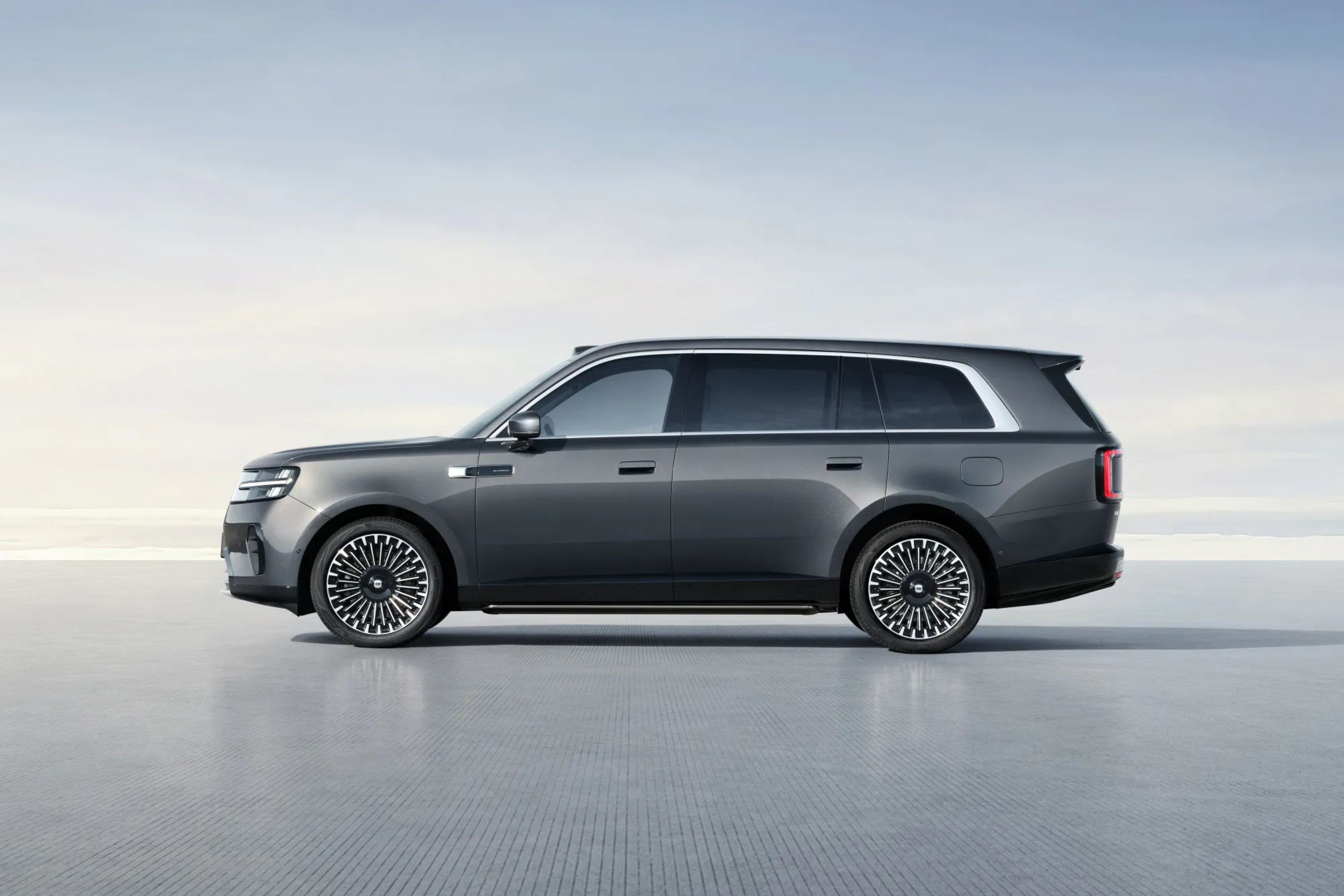 SUV cinza escuro, com rodas de design complexo em preto e prata, parado em uma superfície cinza clara sob um céu azul pálido