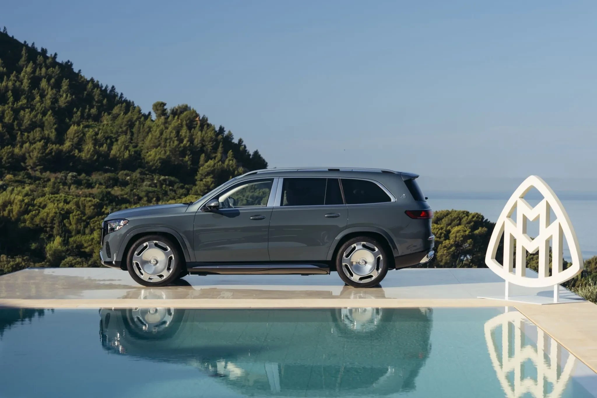 SUV cinza escuro da Maybach estacionado de perfil ao lado de uma piscina, com um grande logo branco da marca à direita. Ao fundo, montanhas verdes e o mar sob céu azul