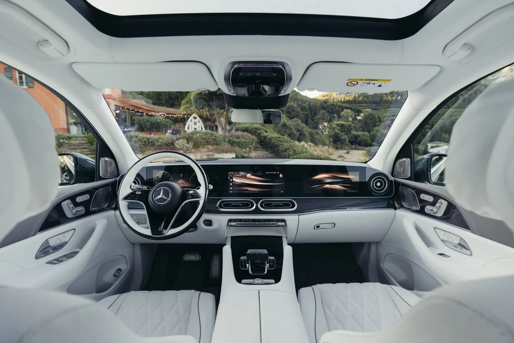 Interior luxuoso de um carro Mercedes-Benz com estofamento branco, painel moderno com telas digitais e volante branco. O teto solar está aberto, revelando uma paisagem verde com montanhas e casas ao fundo