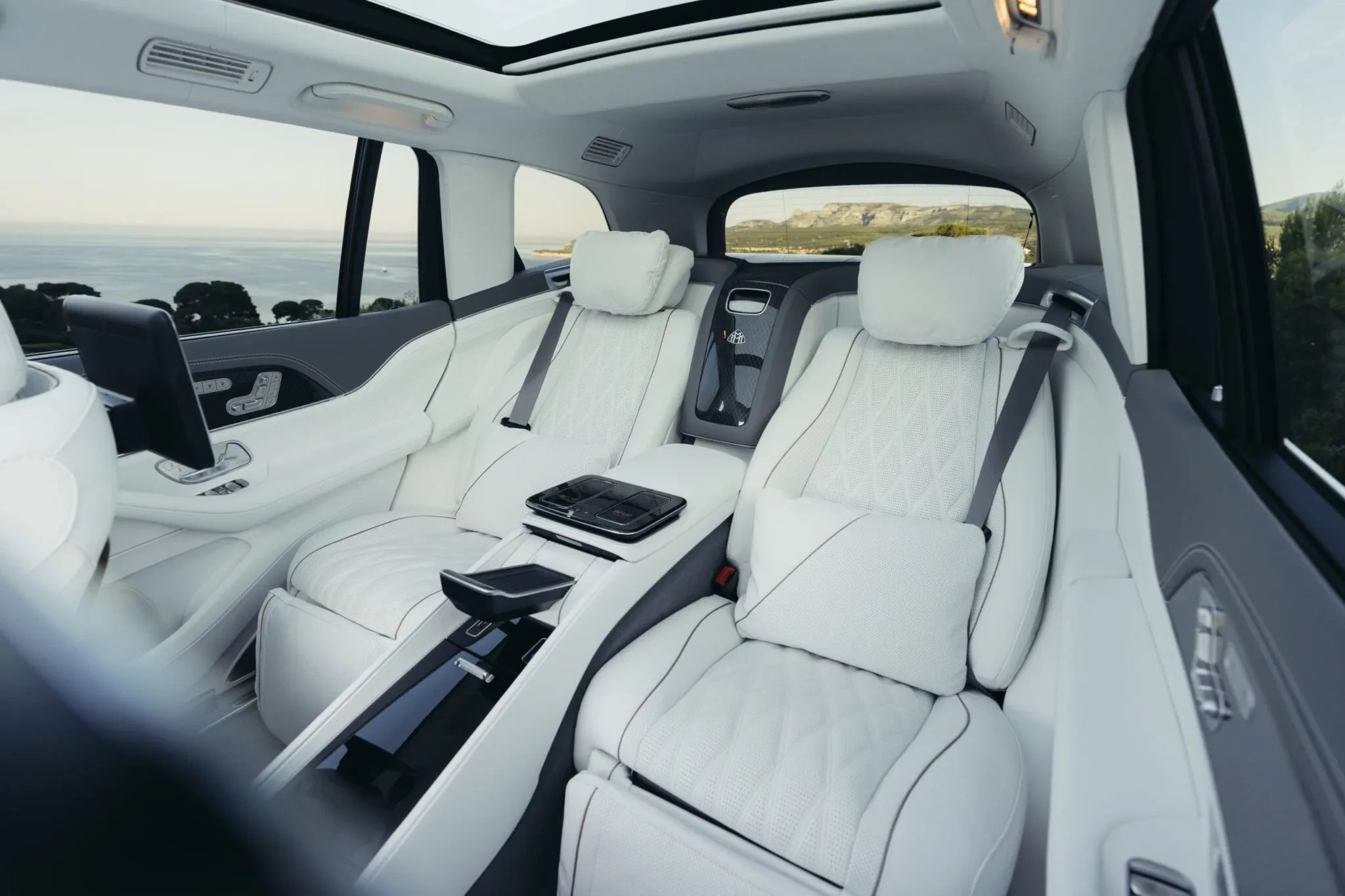 Interior luxuoso de um carro com bancos de couro branco acolchoados, console central com controles e tela, e um tablet acoplado ao banco dianteiro. Ao fundo, uma paisagem costeira com mar e montanhas sob céu claro.