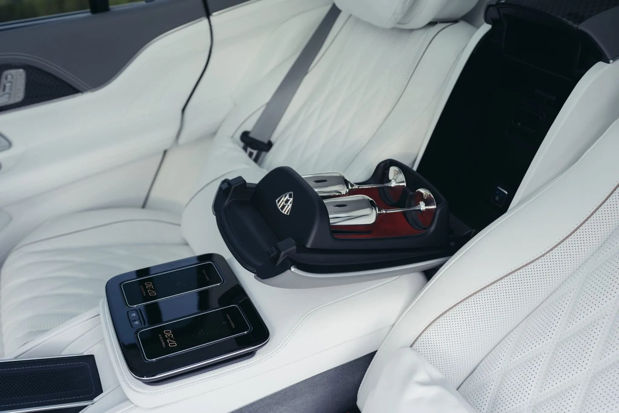 Interior luxuoso de carro com bancos de couro branco, console central com dois smartphones exibindo 07:30 e suporte preto com duas taças de champanhe de metal e base vermelha