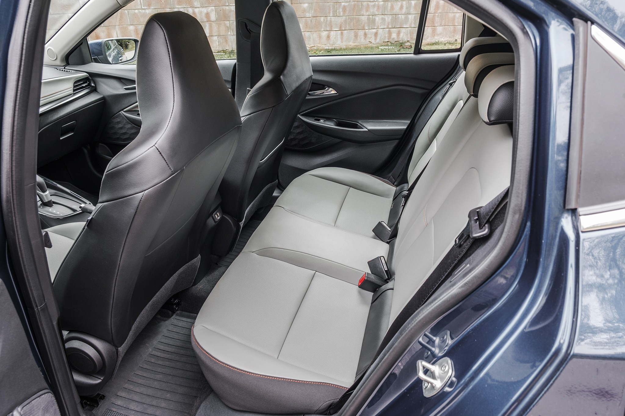 Interior de um carro azul-escuro, mostrando o banco traseiro cinza-claro com detalhes em cinza-escuro e costura vermelha. Os cintos de segurança estão visíveis, e os bancos dianteiros pretos são vistos parcialmente. O assoalho tem tapetes de borracha pretos. A janela traseira revela uma parede de tijolos ao fundo