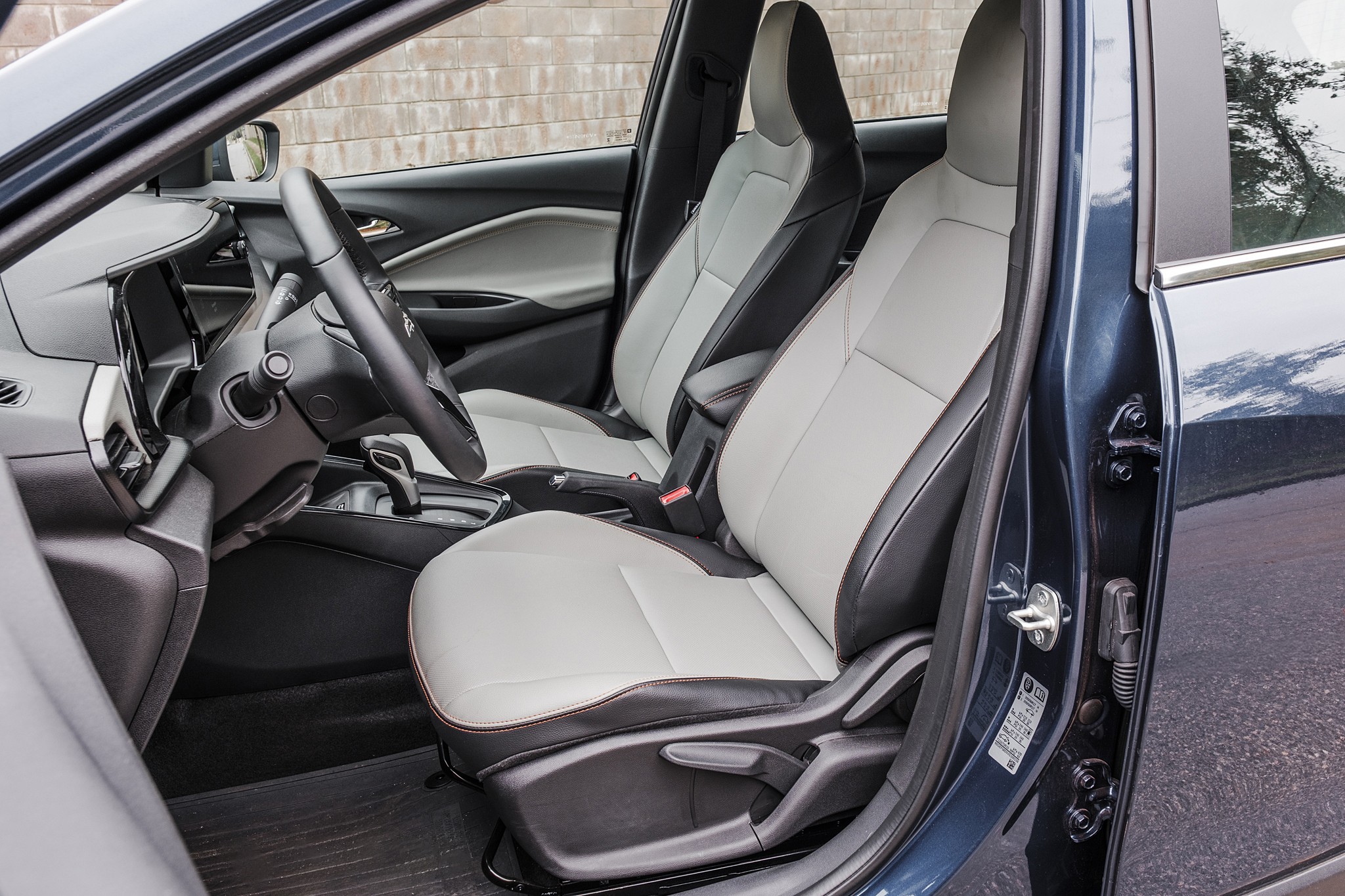 Interior de um carro com bancos dianteiros em couro cinza claro e detalhes pretos, volante escuro e painel com tela central. A porta do motorista está aberta, revelando a lateral do veículo em azul escuro. Ao fundo, uma parede de tijolos claros.