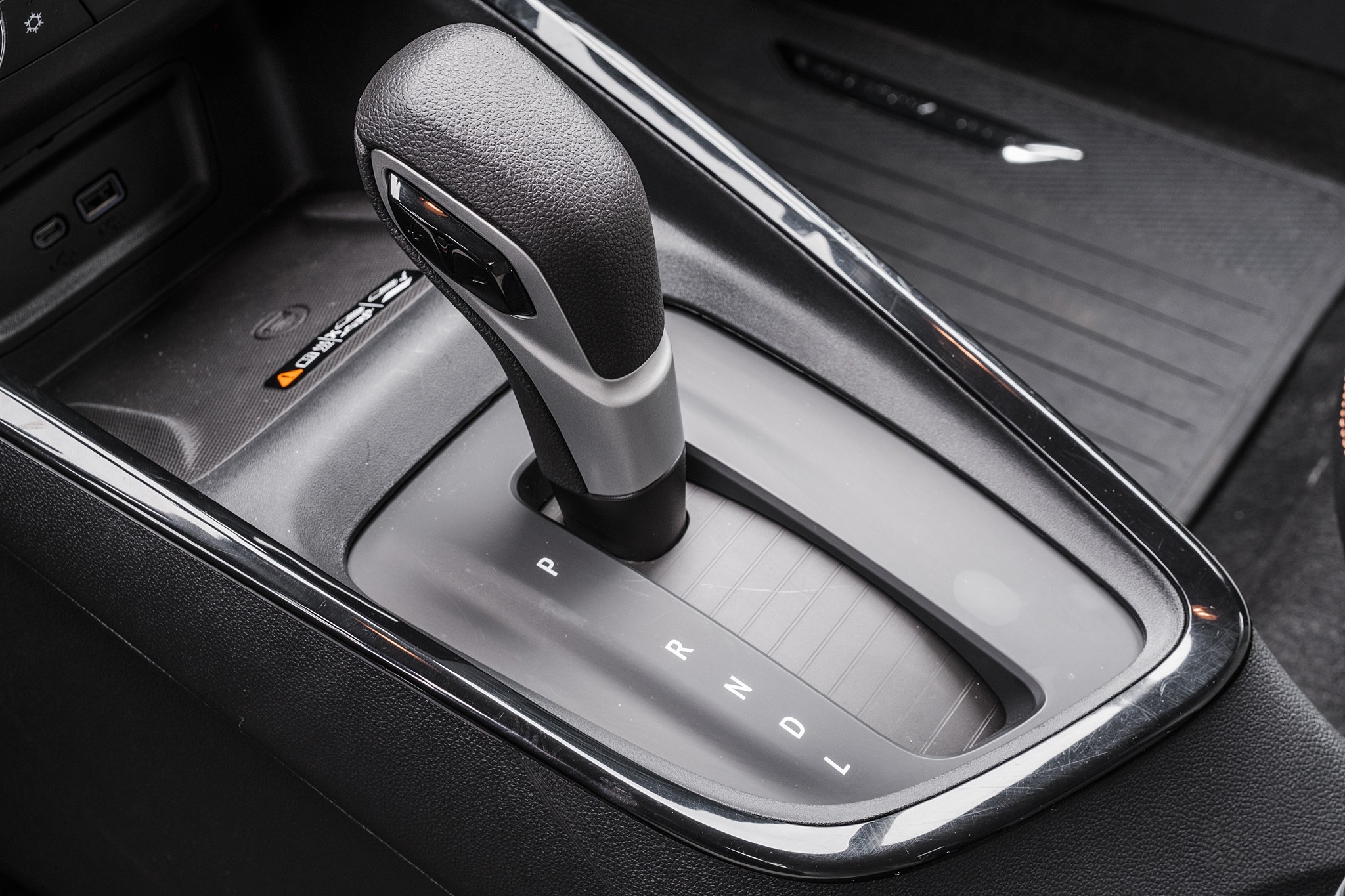 Câmbio automático de um carro, com a alavanca em posição neutra, mostrando as letras P, R, N, D e L. Ao lado, há um compartimento com duas entradas USB e um ícone de carregamento sem fio