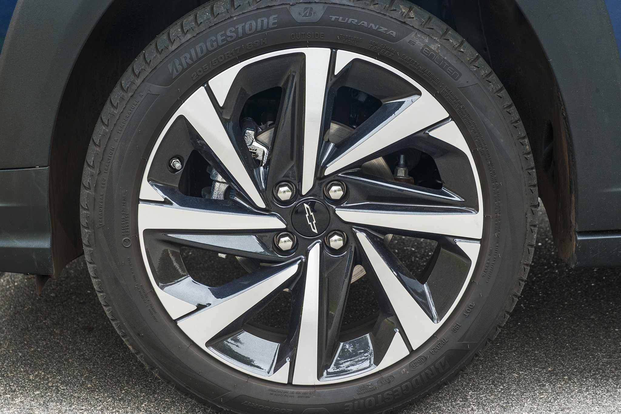 Roda de carro com aro de liga leve preto e prata, pneu Bridgestone Turanza com banda de rodagem visível, e o emblema da Chevrolet no centro. O para-lama escuro do veículo aparece na parte superior, e o asfalto cinza na parte inferior