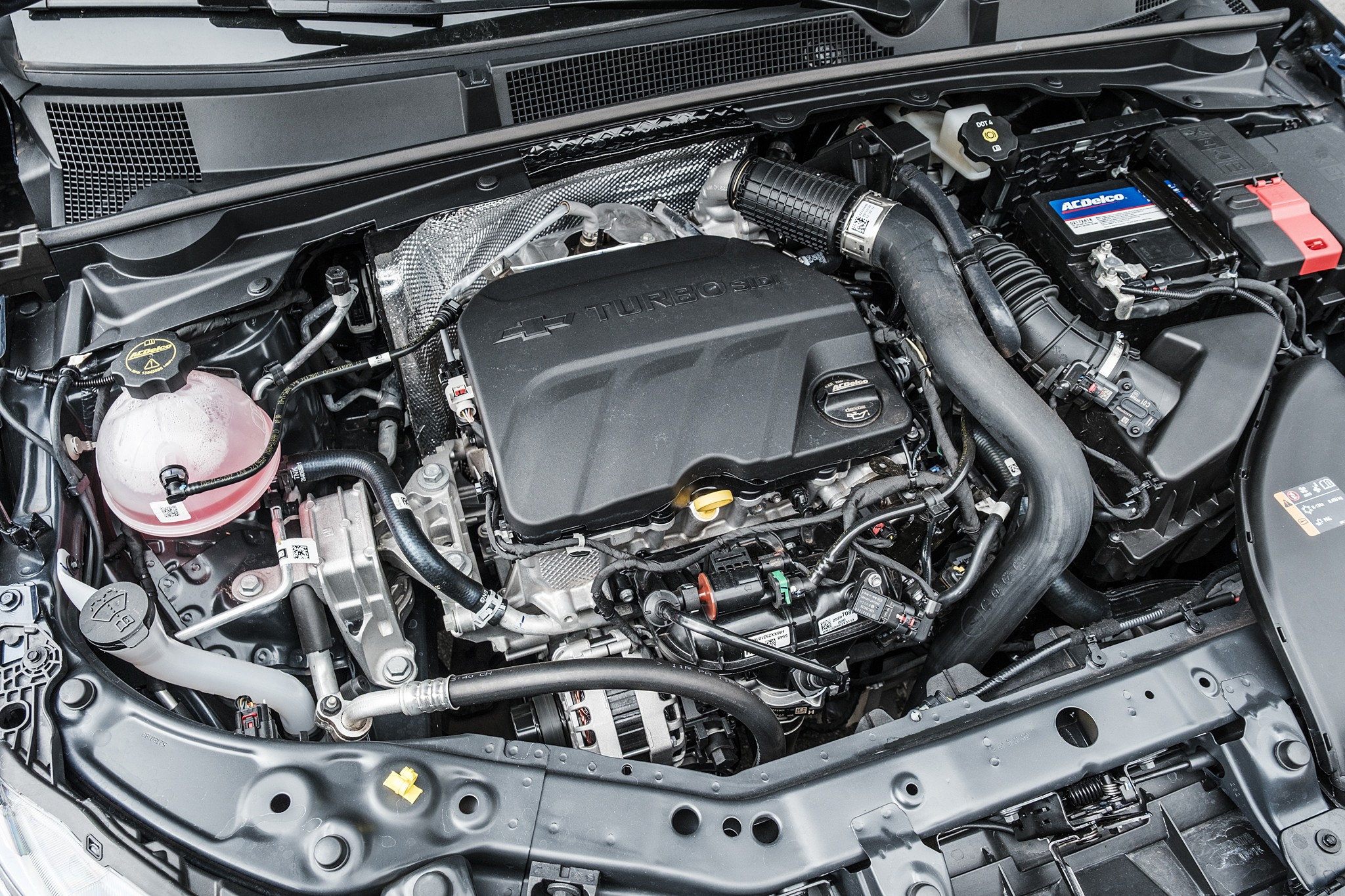 Motor de carro moderno, com a tampa do motor preta em destaque, mangueiras, fios e reservatórios visíveis, incluindo um reservatório de fluido rosa translúcido e uma bateria ACDelco azul