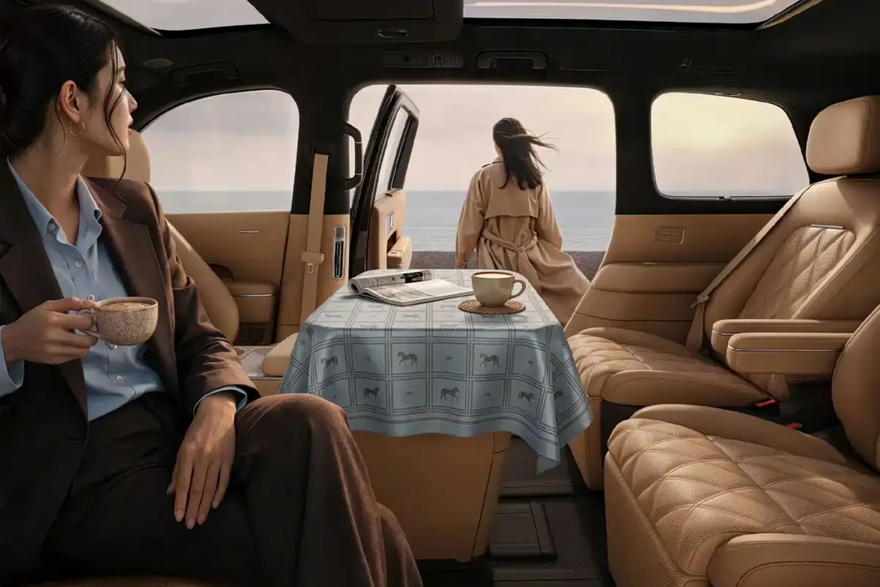 Mulher de terno marrom e camisa azul sentada em carro de luxo, segurando xícara de café, olhando para outra mulher de casaco bege em frente ao mar. Uma mesa com toalha xadrez e xícara de café está entre elas