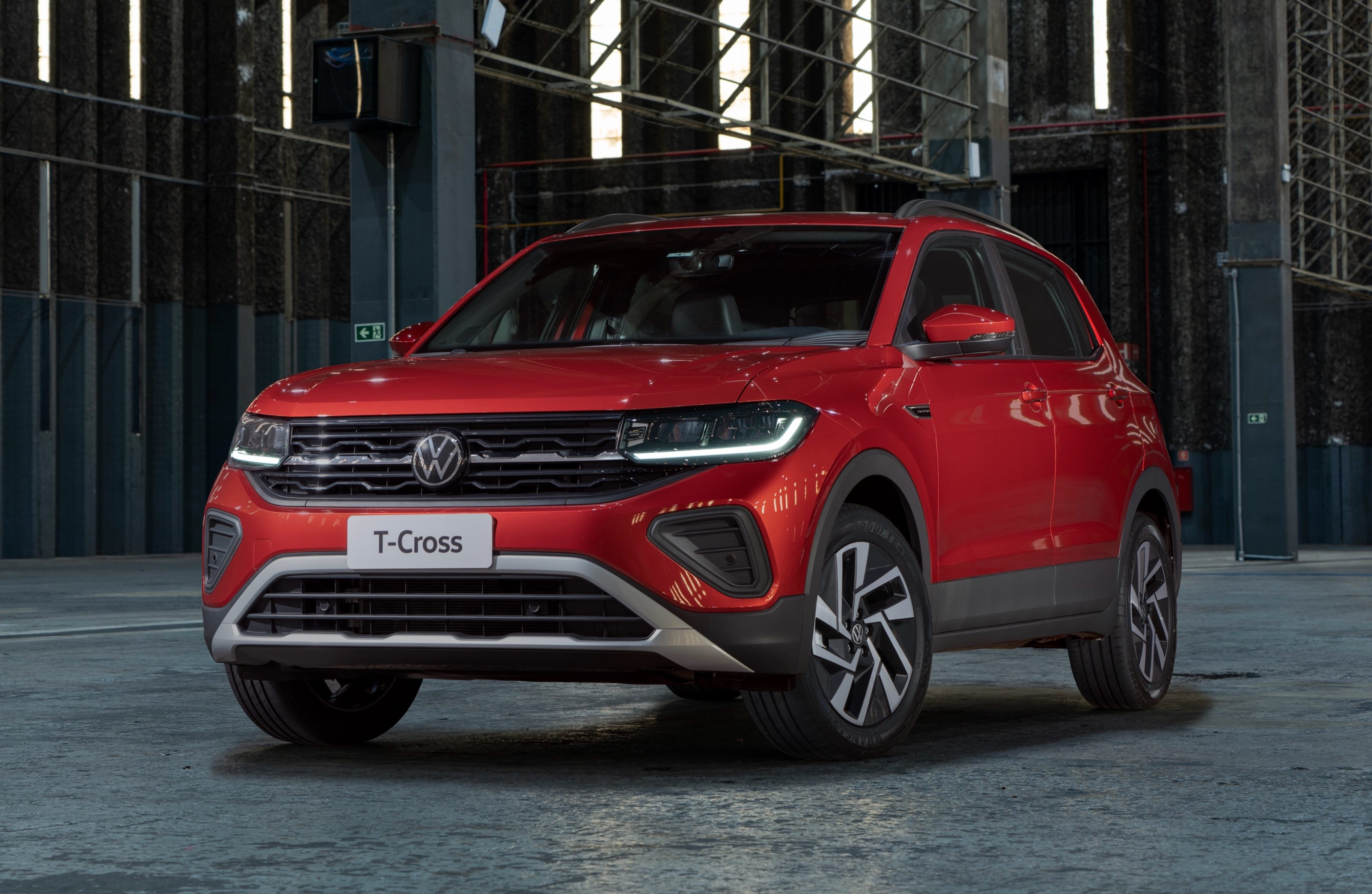 SUV Volkswagen T-Cross vermelho vibrante, com faróis e luzes diurnas acesas, estacionado em um galpão industrial escuro com paredes texturizadas e iluminação vertical