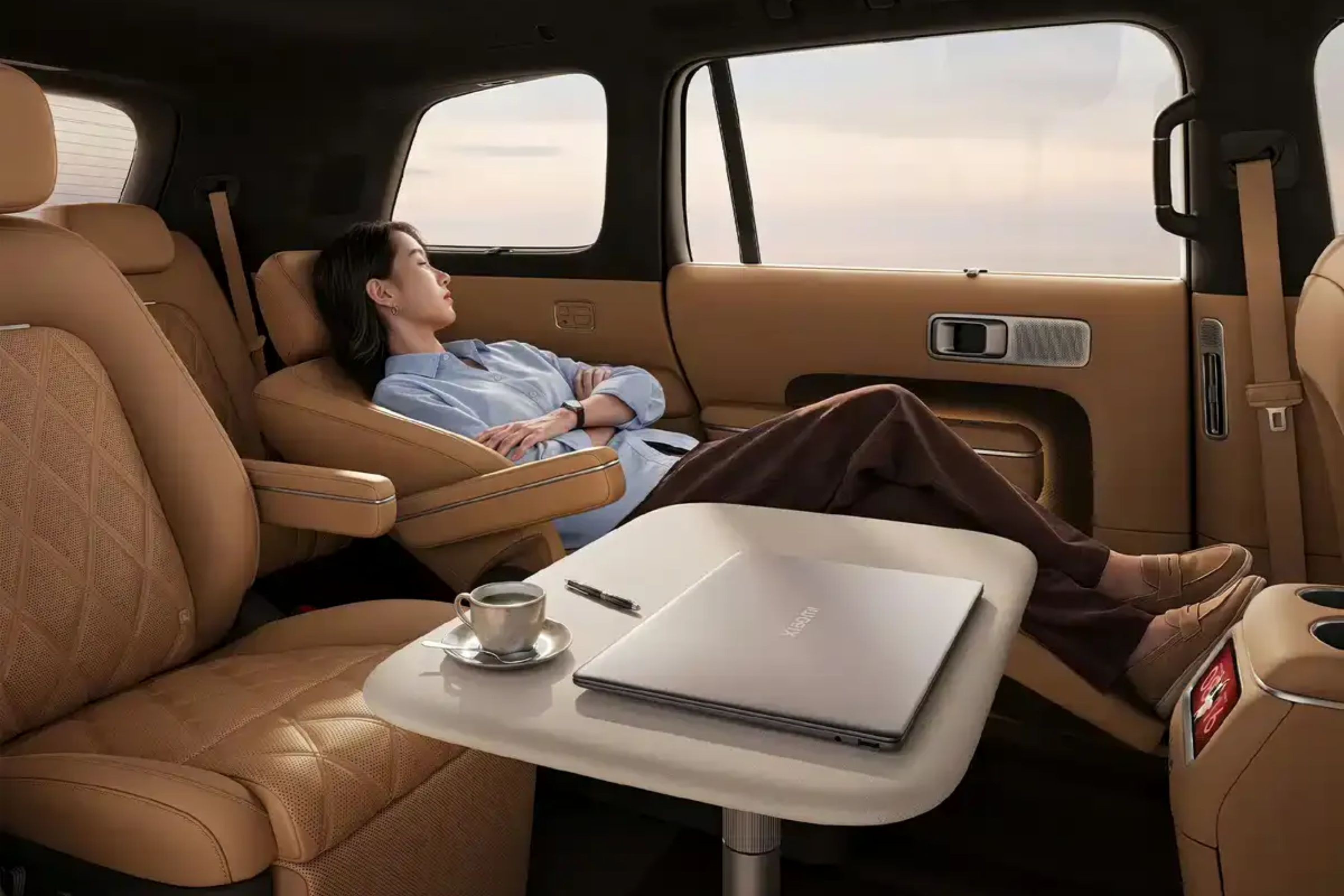 Mulher asiática de camisa azul e calça marrom relaxa em assento reclinável de carro luxuoso, com os olhos fechados. Uma mesa lateral com notebook prateado, caneta e xícara de café está à sua frente. O interior do veículo é bege e marrom, com janelas mostrando um céu claro ao fundo