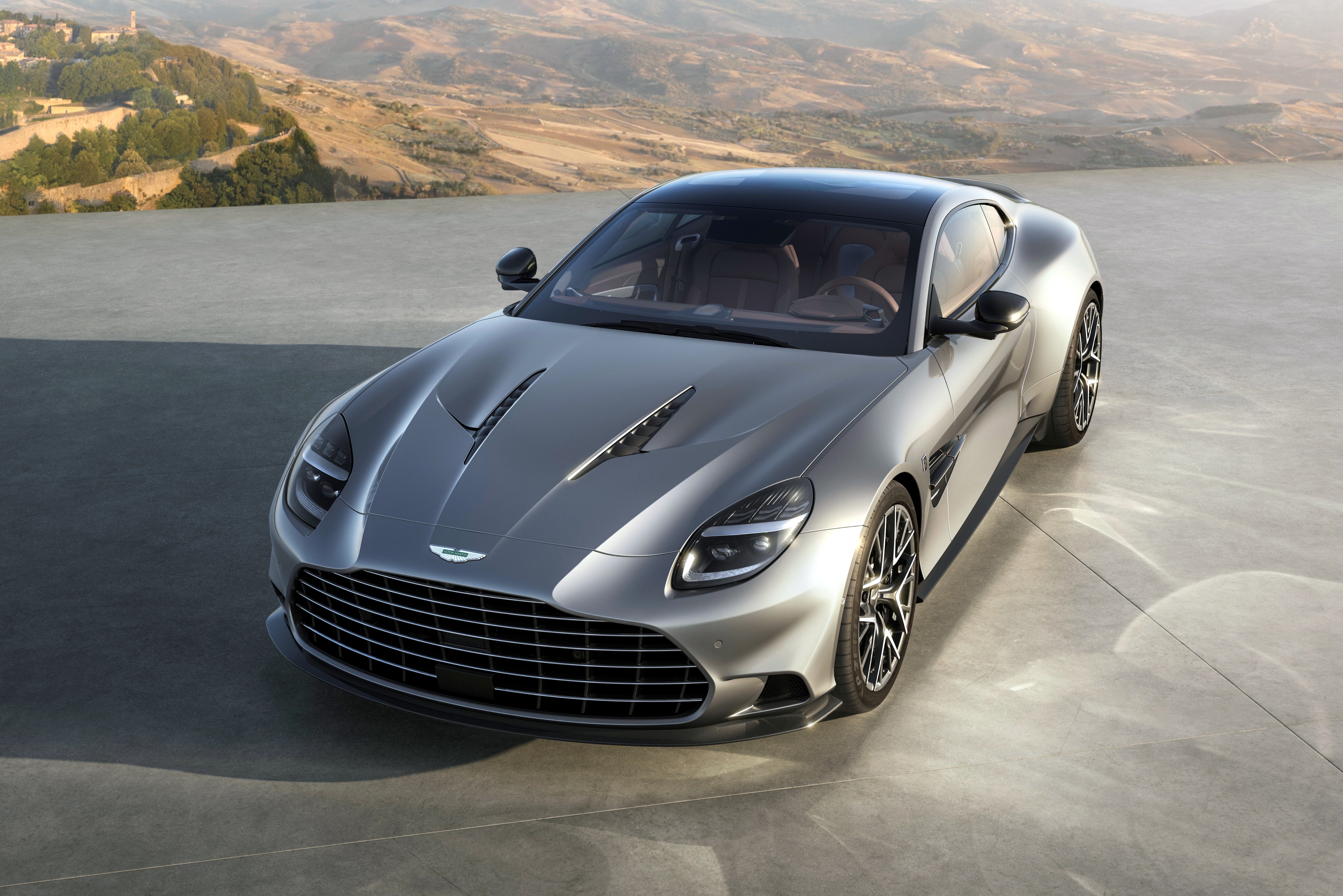 Carro esportivo Aston Martin prateado com teto preto, estacionado em uma superfície cinza. Ao fundo, montanhas e um vilarejo distante sob um céu claro