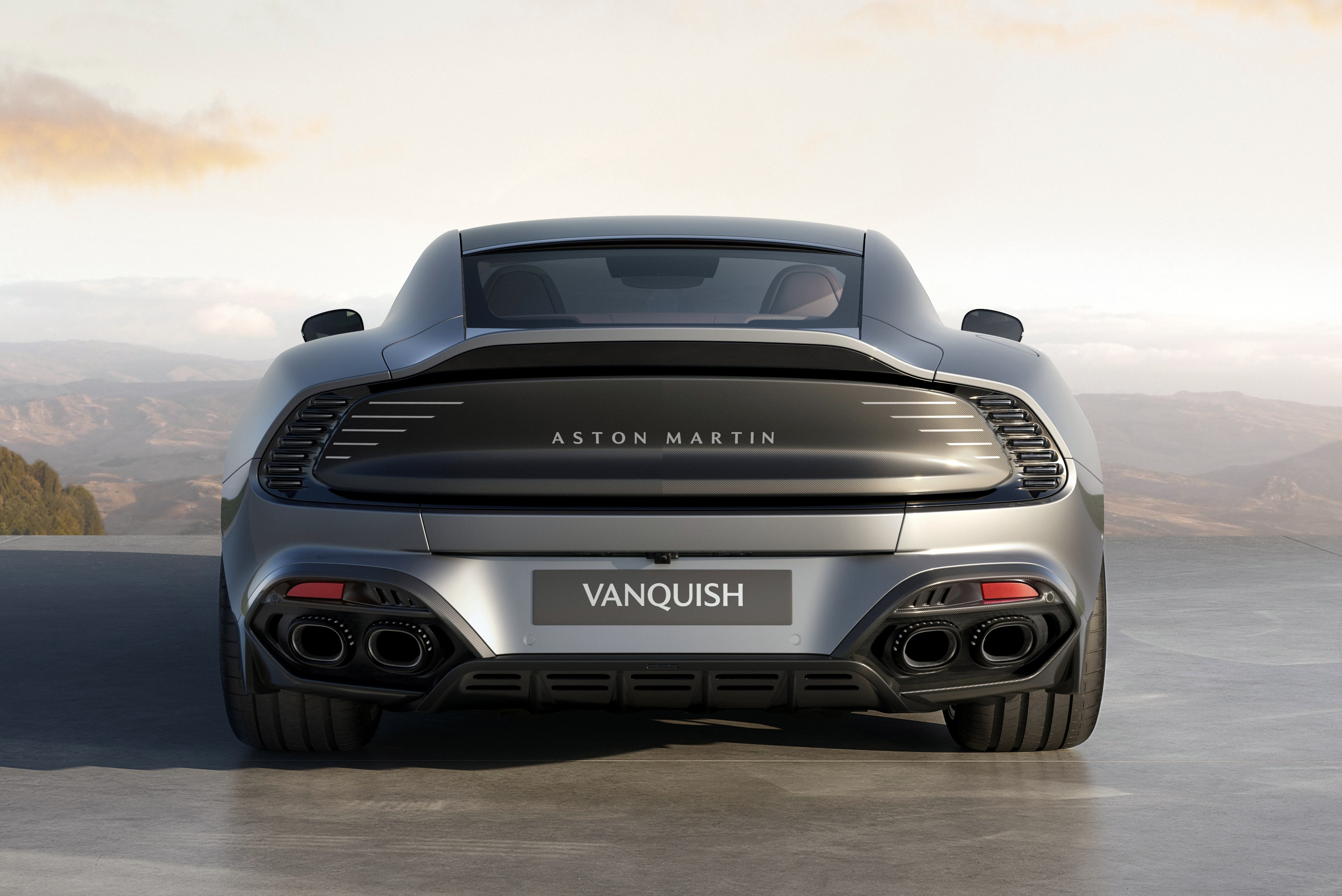 Traseira de um carro esportivo Aston Martin Vanquish cinza metálico, com quatro saídas de escapamento pretas e lanternas vermelhas, estacionado em uma estrada com montanhas ao fundo e céu claro