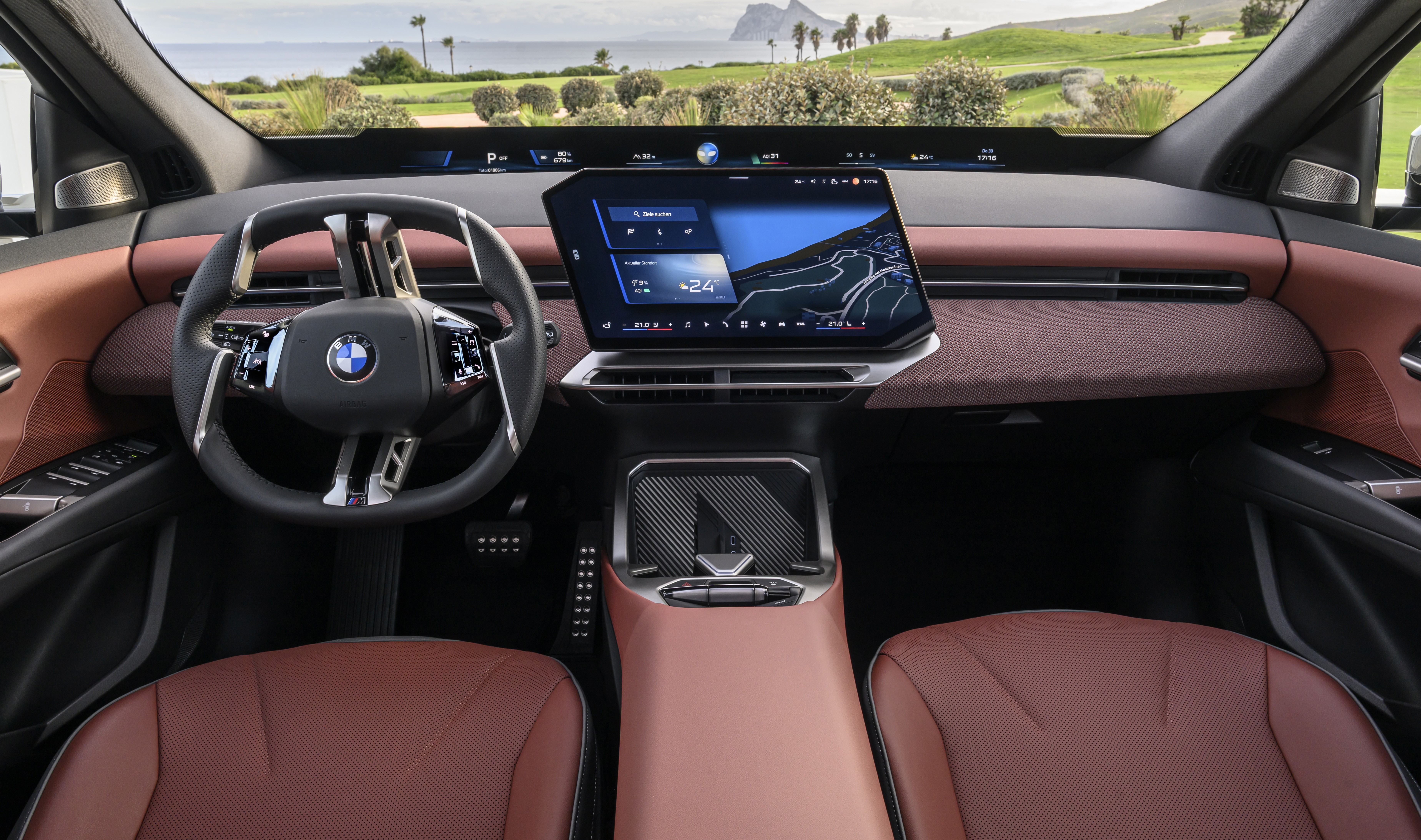 Interior de um carro moderno com volante BMW, painel marrom e tela central exibindo mapa e temperatura. Bancos de couro marrom e paisagem verde com mar ao fundo visível pelo para-brisa