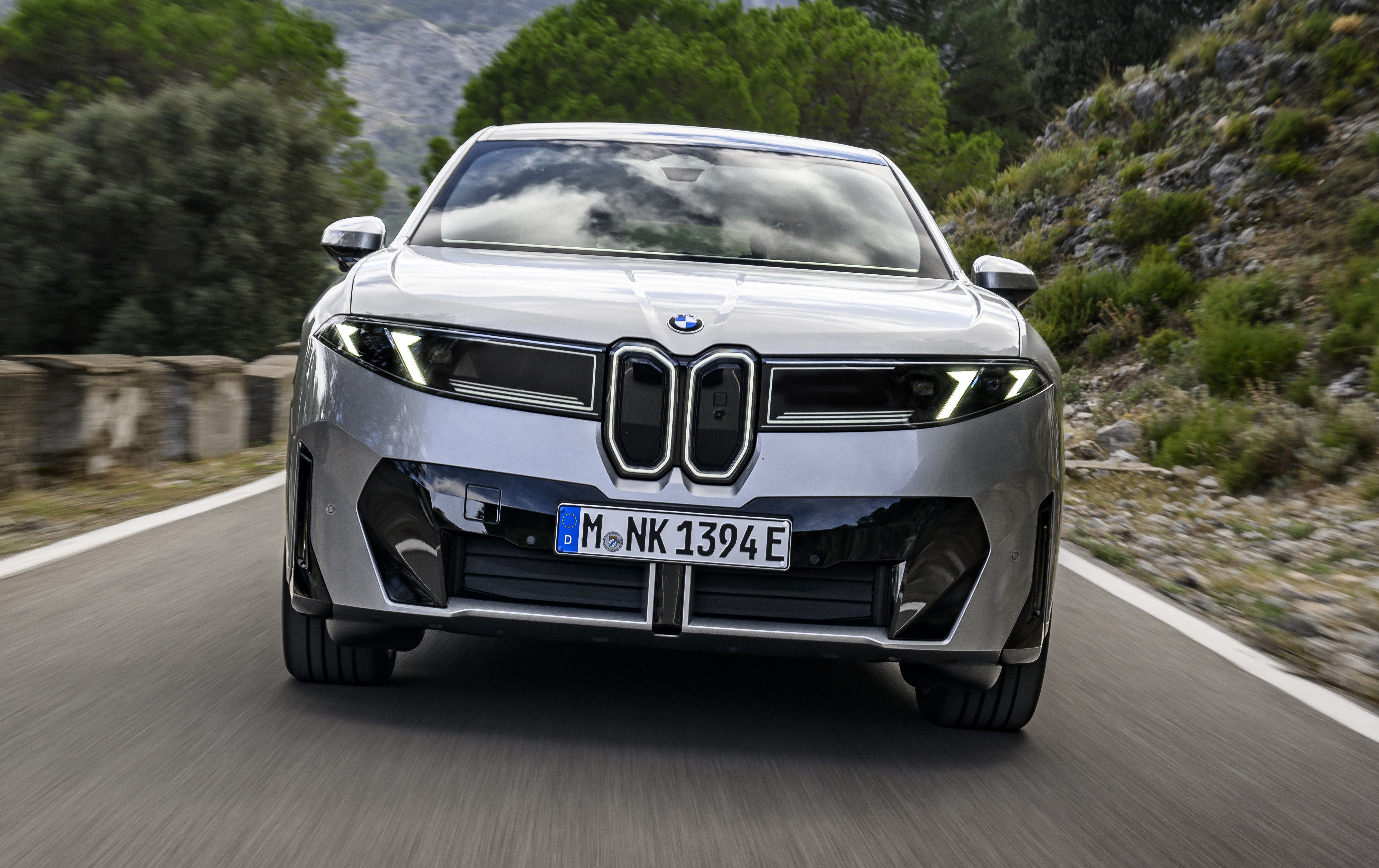Carro BMW prateado em movimento, visto de frente, com faróis acesos e grade dupla em destaque, em uma estrada sinuosa com vegetação e montanhas ao fundo