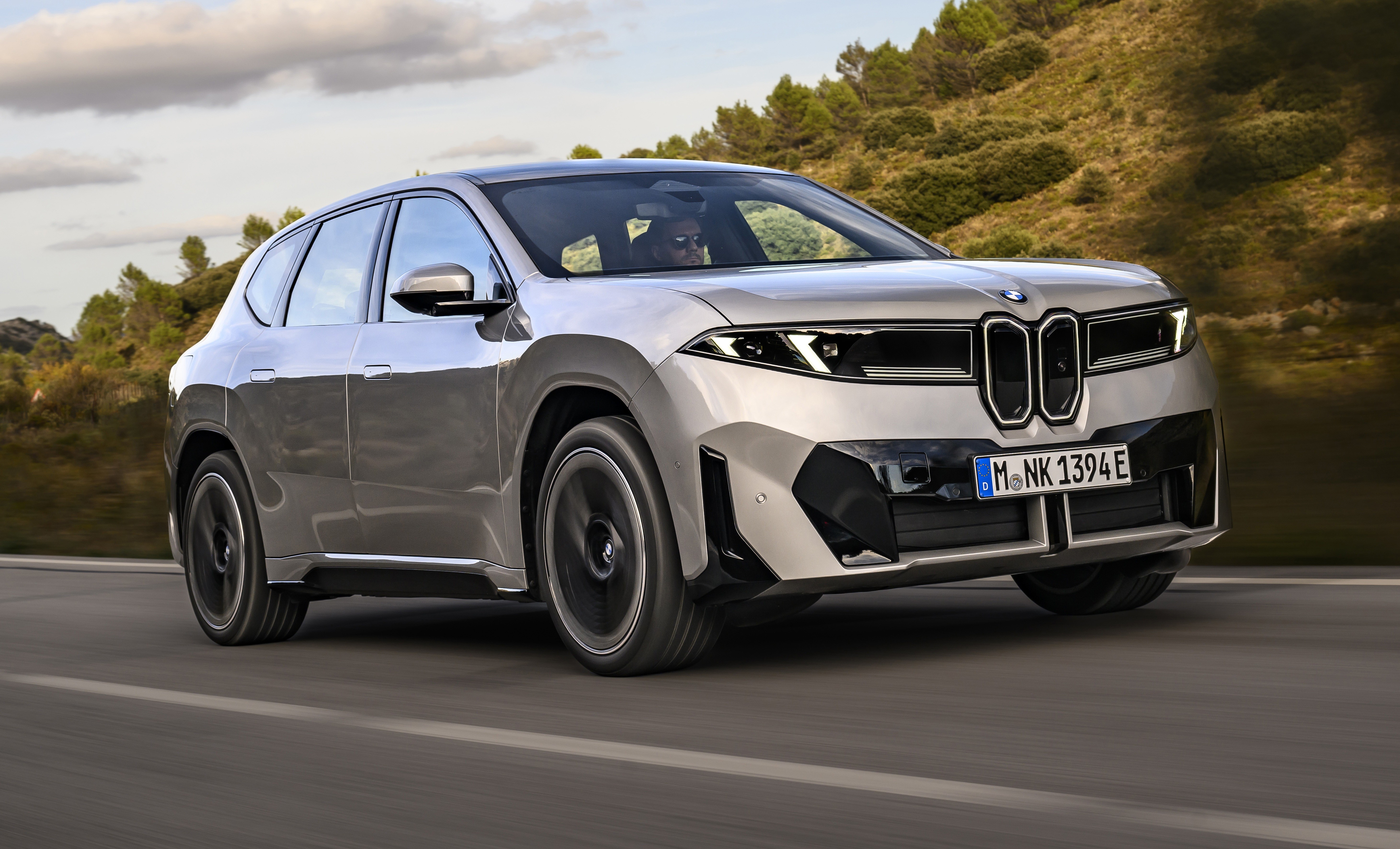 SUV BMW iX prateado em movimento rápido numa estrada, com montanhas e céu nublado ao fundo