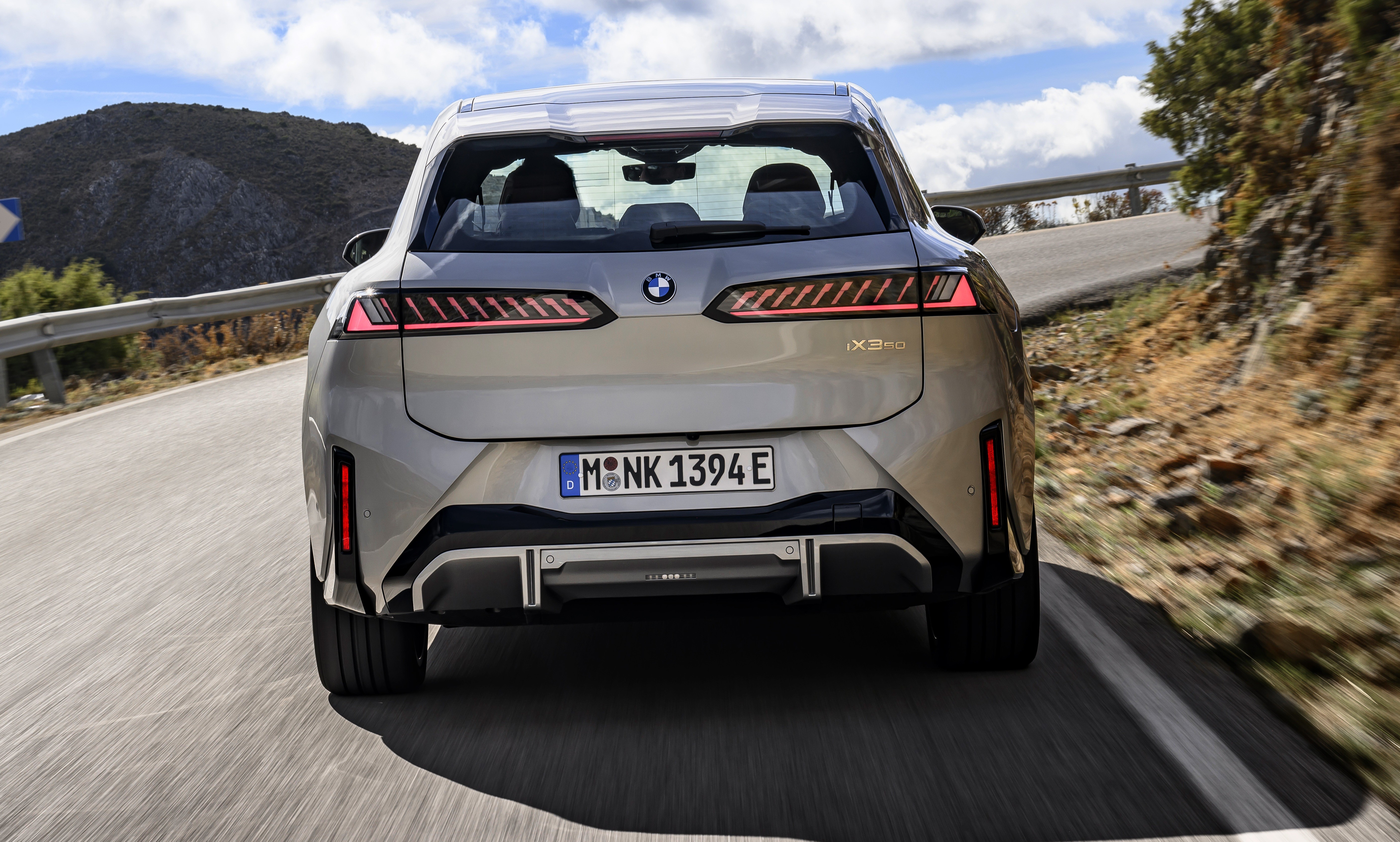 Traseira de um carro BMW iX3 elétrico cinza claro em uma estrada sinuosa, com montanhas e céu azul com nuvens ao fundo