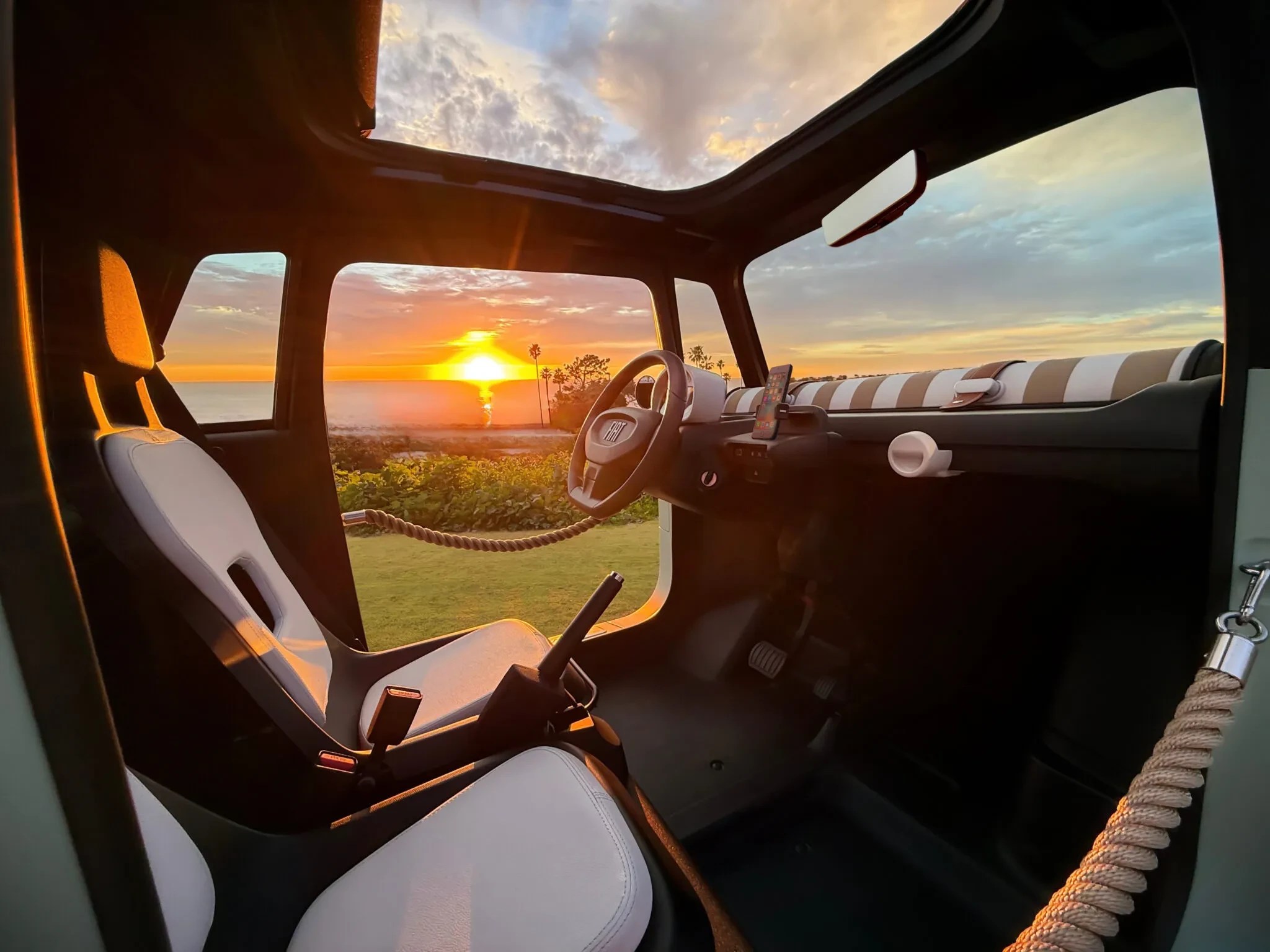 Interior de um carro compacto com teto solar aberto, bancos brancos e painel listrado em bege e branco. O sol se põe no horizonte do mar, visível pela janela, iluminando o interior do veículo com tons alaranjados. Palmeiras e vegetação verde aparecem à distância