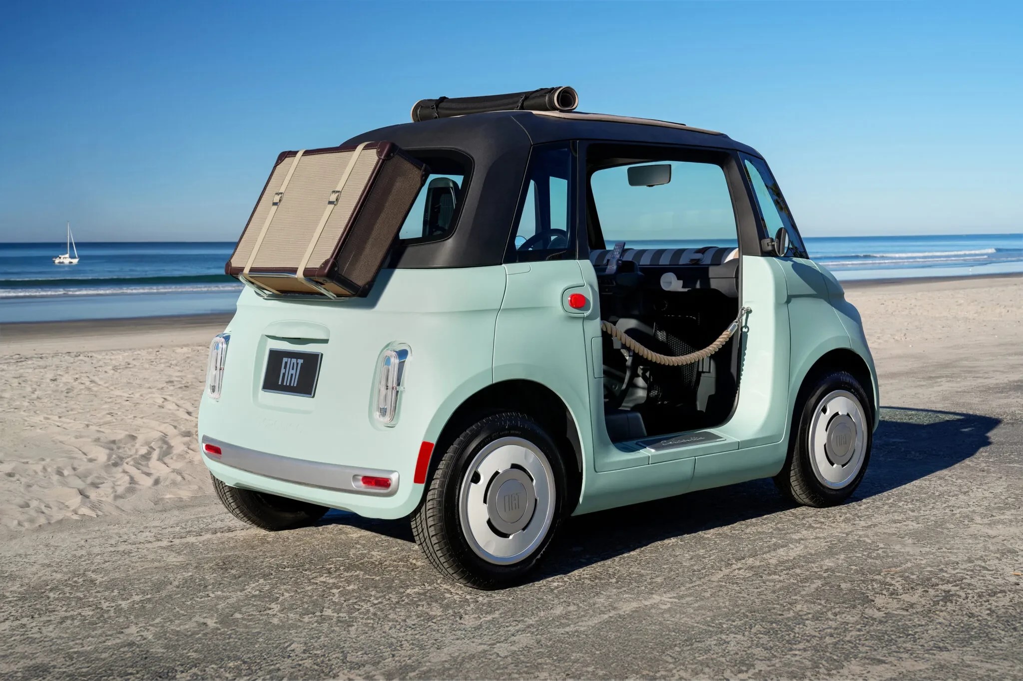 Carro Fiat 500 elétrico azul-claro, com teto preto e porta aberta, estacionado na areia da praia. Uma mala bege está presa na traseira e um veleiro branco navega no mar azul ao fundo