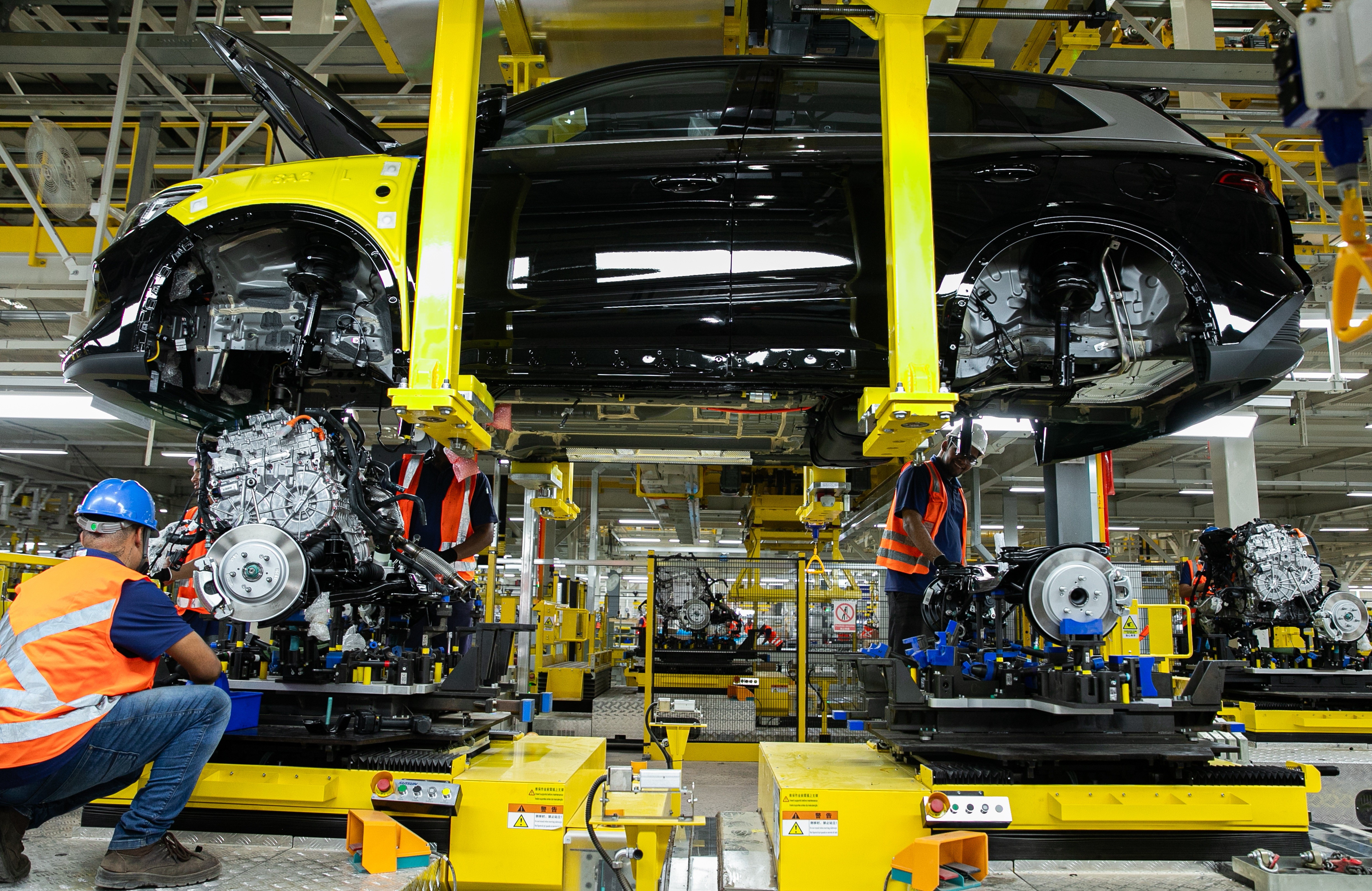 Homens em coletes laranja e capacetes azuis trabalham na montagem de carros em uma linha de produção industrial. Um carro preto suspenso por guindastes amarelos tem o capô aberto, enquanto os trabalhadores instalam motores e componentes em chassis. O ambiente é iluminado e repleto de equipamentos e máquinas amarelas