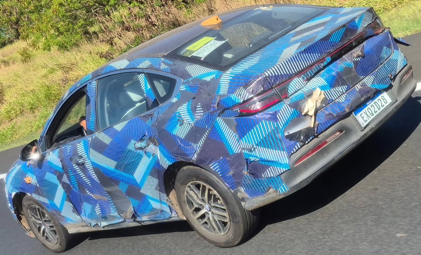 Carro camuflado com adesivos azuis e pretos em padrão geométrico, visto de traseira e lateral, em uma estrada com vegetação ao fundo. A placa traseira é EXQ2D28