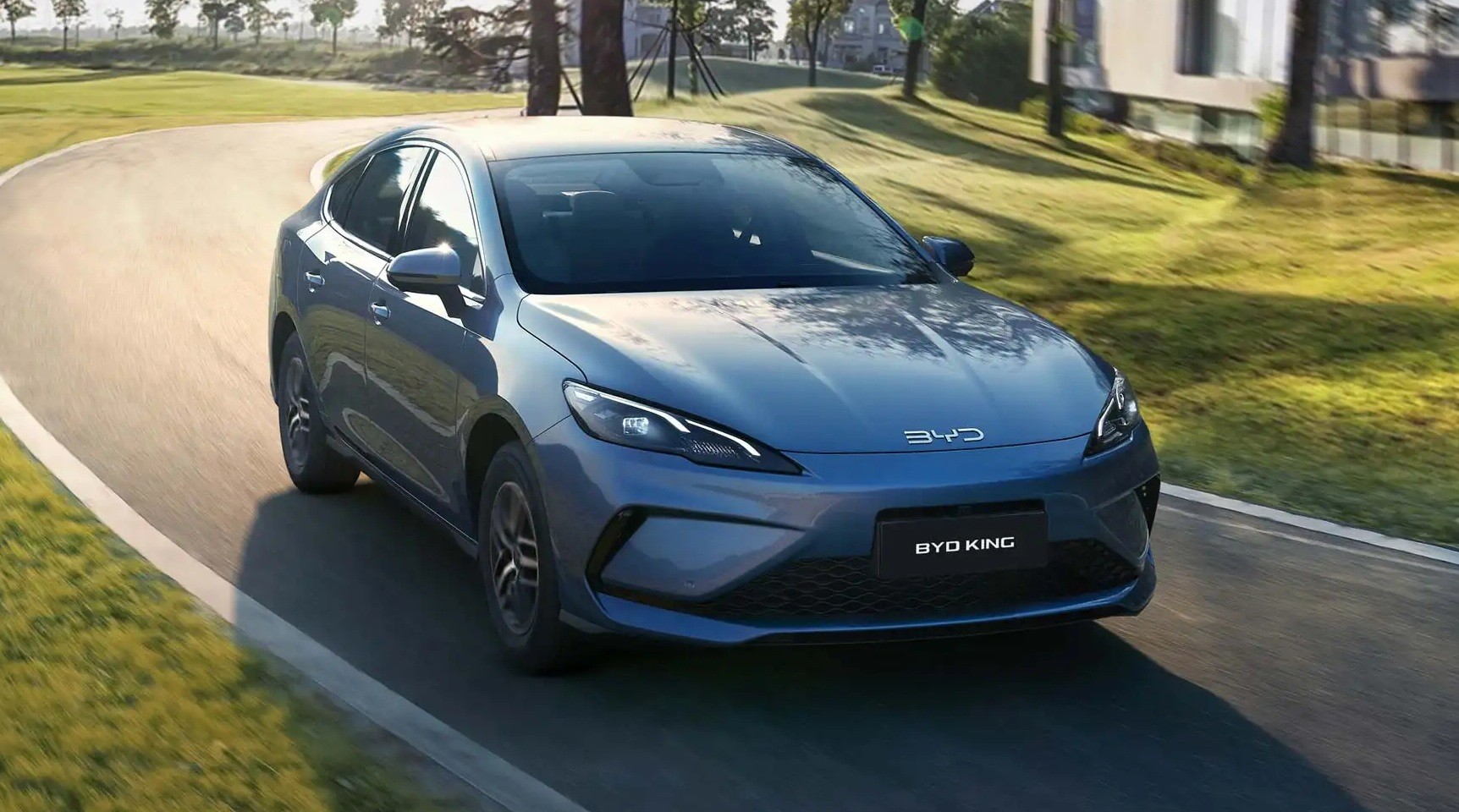 Sedan BYD King azul metálico em uma curva suave de estrada asfaltada, com grama verde e árvores ao fundo, sob luz solar
