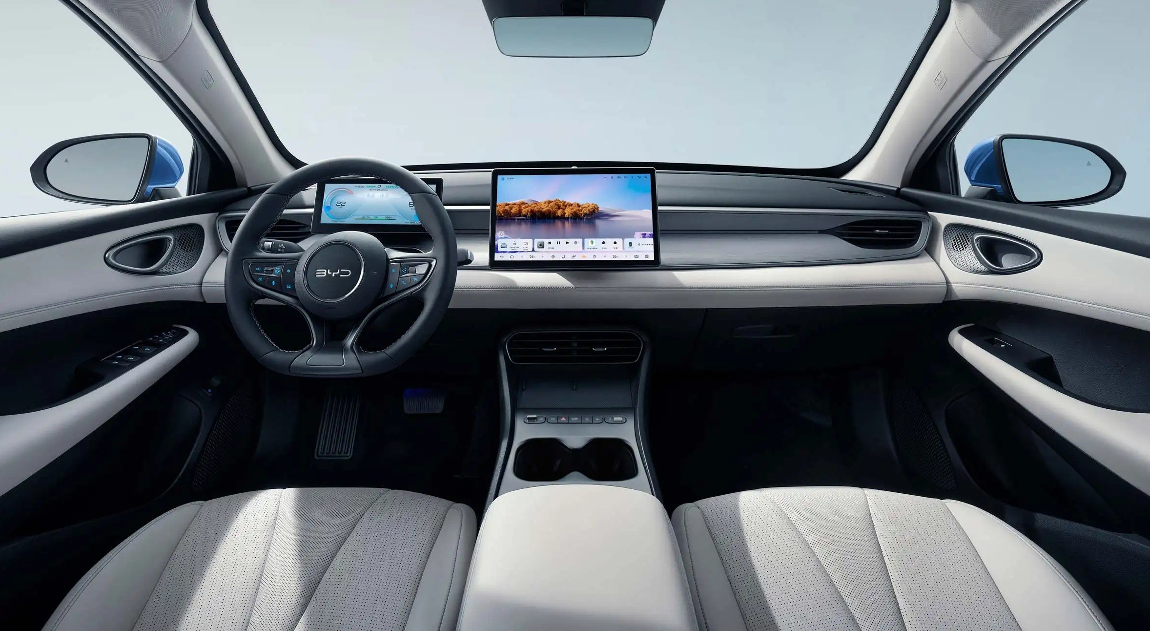 Interior de um carro moderno com bancos claros, volante multifuncional escuro e uma grande tela central exibindo uma paisagem com árvores e água. O painel é cinza claro e há um console central com porta-copos.