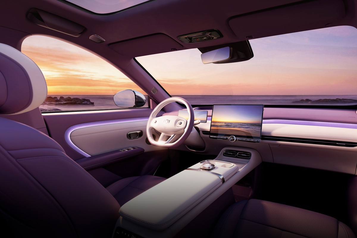 Interior de carro moderno com bancos roxos e detalhes brancos, volante branco e tela multimídia exibindo pôr do sol na praia. O painel tem iluminação ambiente roxa, e o teto solar panorâmico reflete o céu alaranjado sobre o oceano com rochas à distância