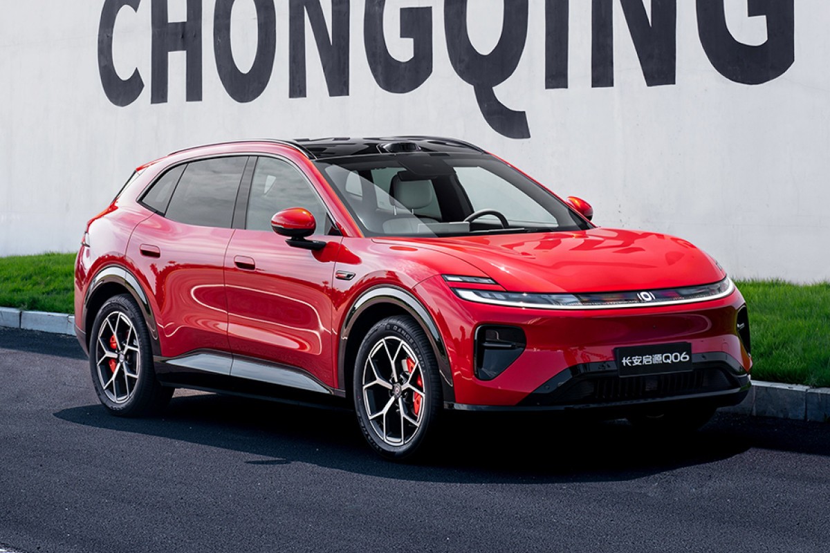 SUV vermelho brilhante, modelo Changan Q06, estacionado em asfalto escuro com uma parede branca e grama verde ao fundo. O carro tem teto preto, rodas esportivas e faróis de LED finos. O nome CHONGQING está escrito em letras grandes e pretas na parede