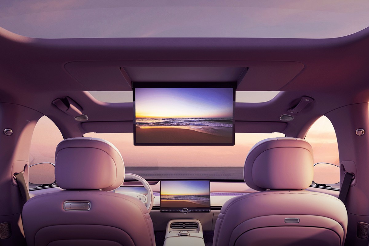 Interior de carro moderno com bancos roxos claros, teto solar e duas telas exibindo um pôr do sol na praia. Uma tela grande desce do teto, e outra menor está no painel central. O volante branco é visível à esquerda, e o céu rosado é visto pelas janelas