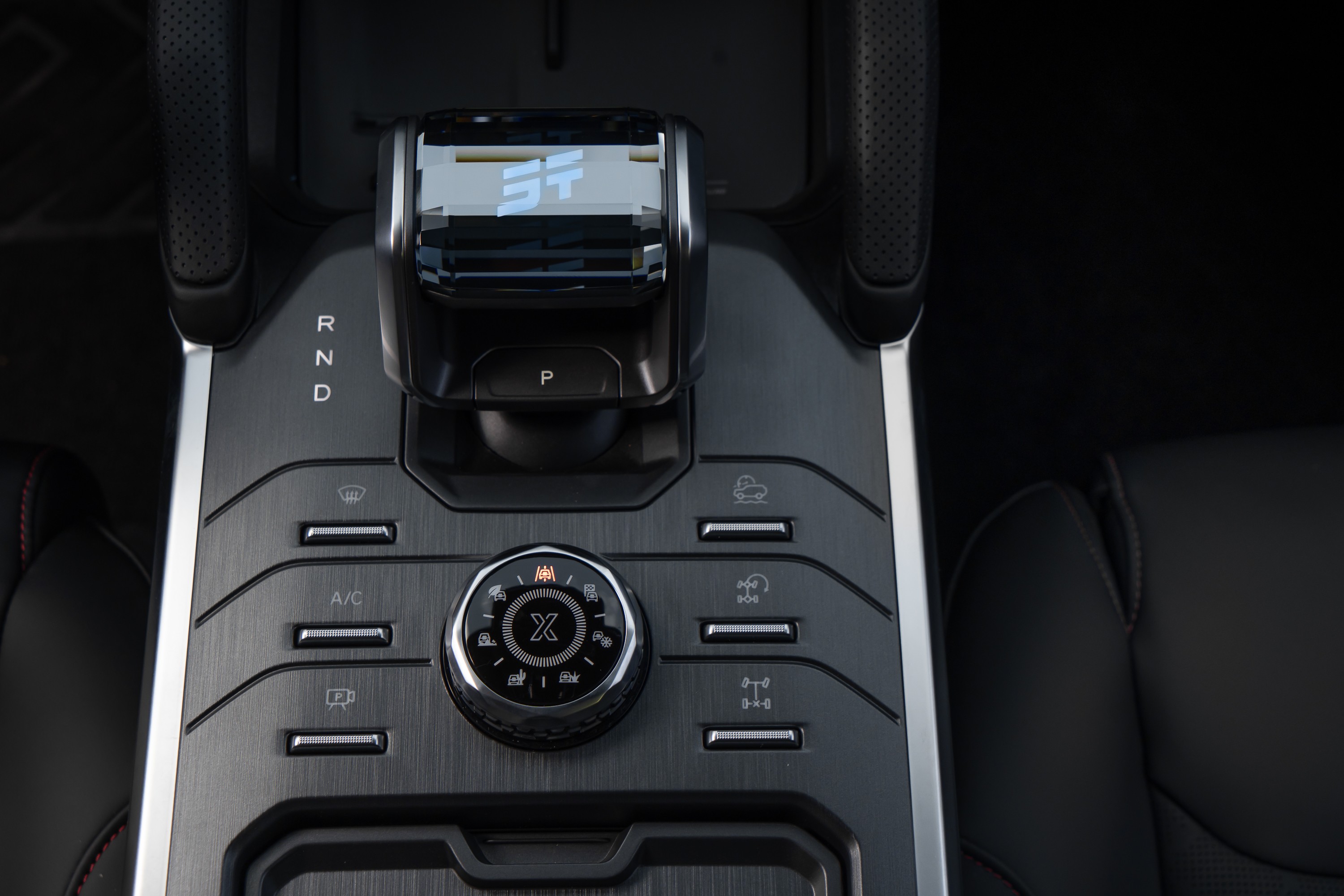 Console central de um carro com câmbio automático, botões de controle do ar-condicionado, ventilação e tração, e um seletor giratório com um X no centro, em tons de cinza e preto