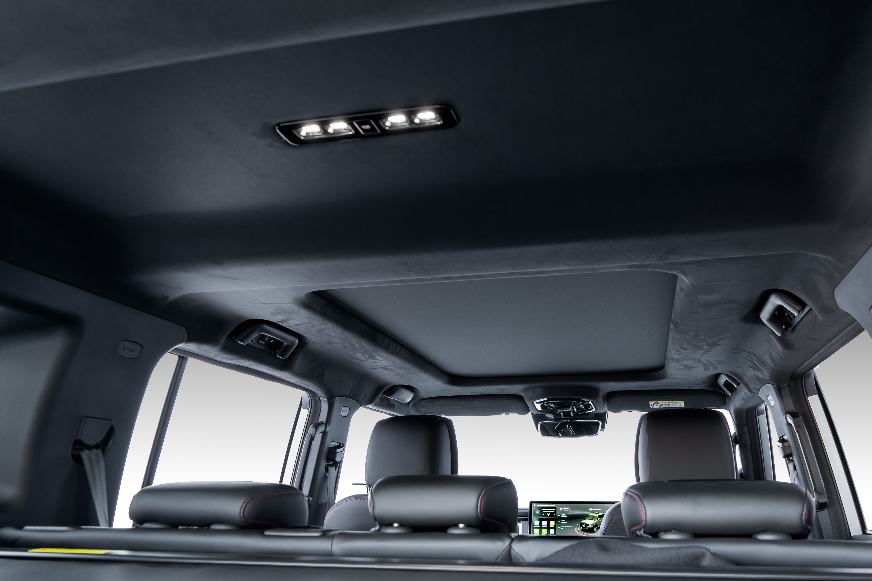 Interior de um carro de luxo com teto solar panorâmico e forro escuro. Luzes de LED acesas no teto, bancos de couro pretos com costura vermelha e uma tela multimídia visível no banco traseiro. O ambiente é sofisticado e moderno