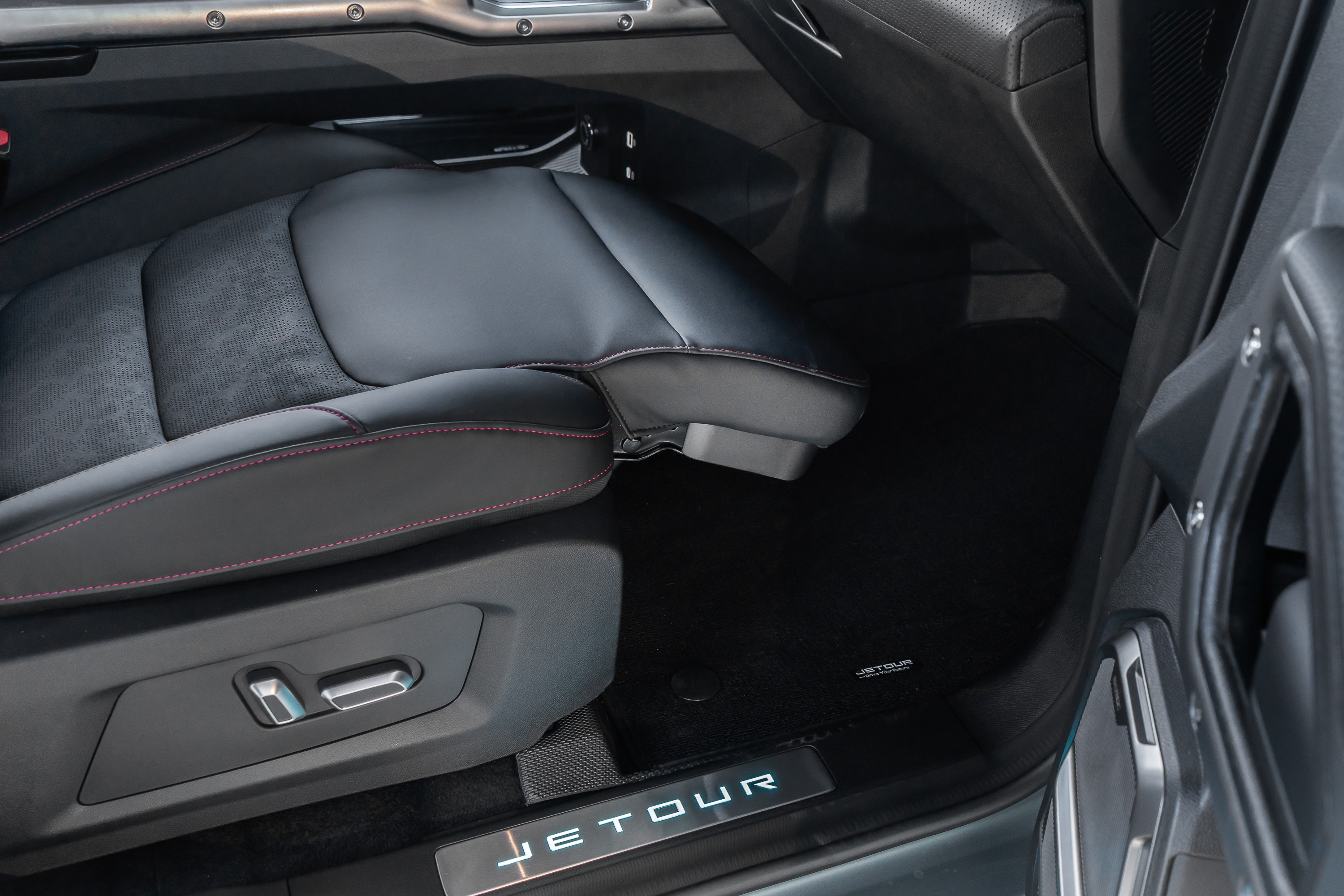 Interior de carro com banco de couro preto e costura vermelha, ajuste elétrico visível na lateral. O tapete escuro tem o logo JETOUR e a soleira da porta exibe o nome JETOUR iluminado em azul claro