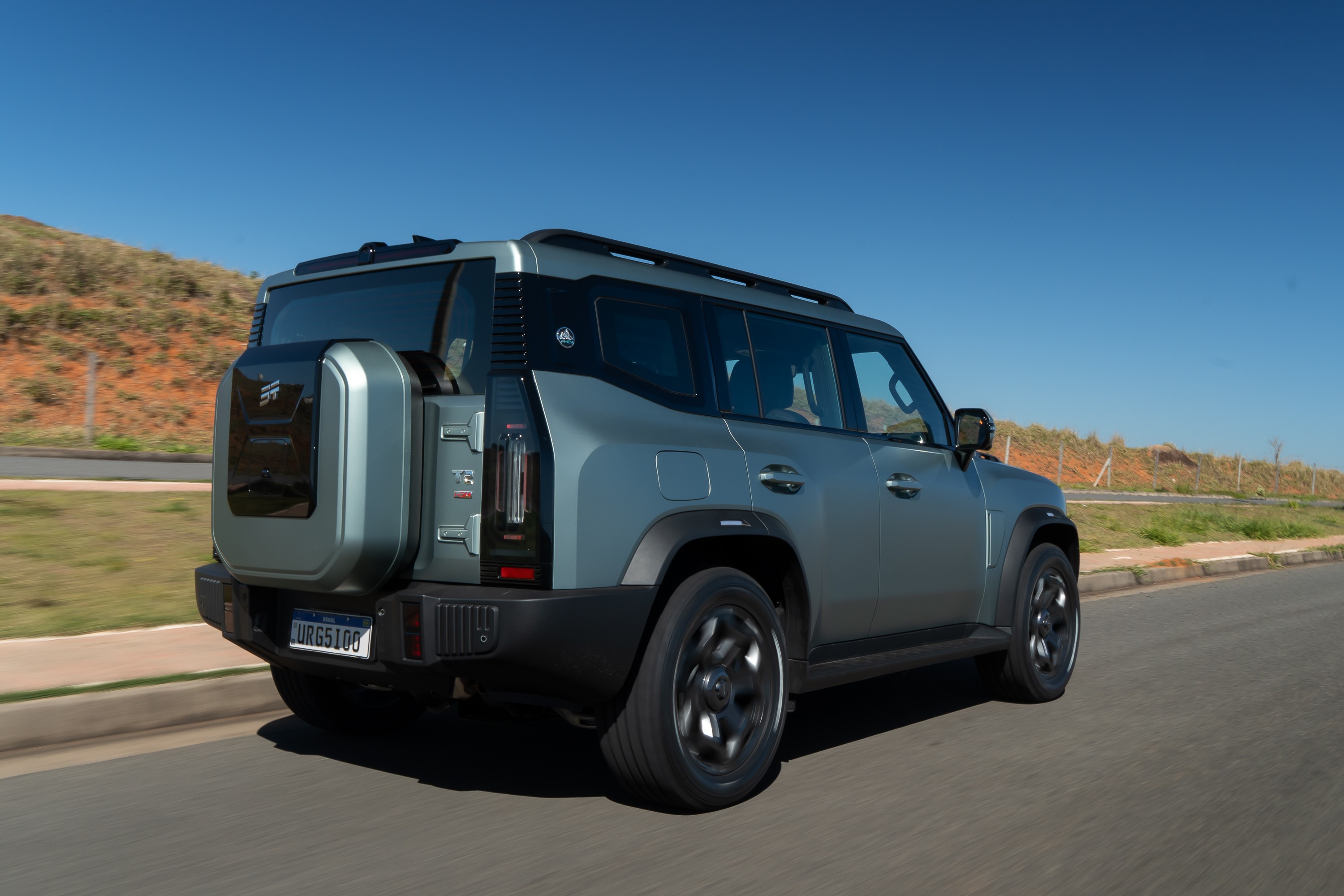 SUV cinza-azulado com estepe na traseira, rodando em uma estrada asfaltada sob céu azul claro. O veículo tem rack de teto e detalhes pretos, com uma colina marrom ao fundo
