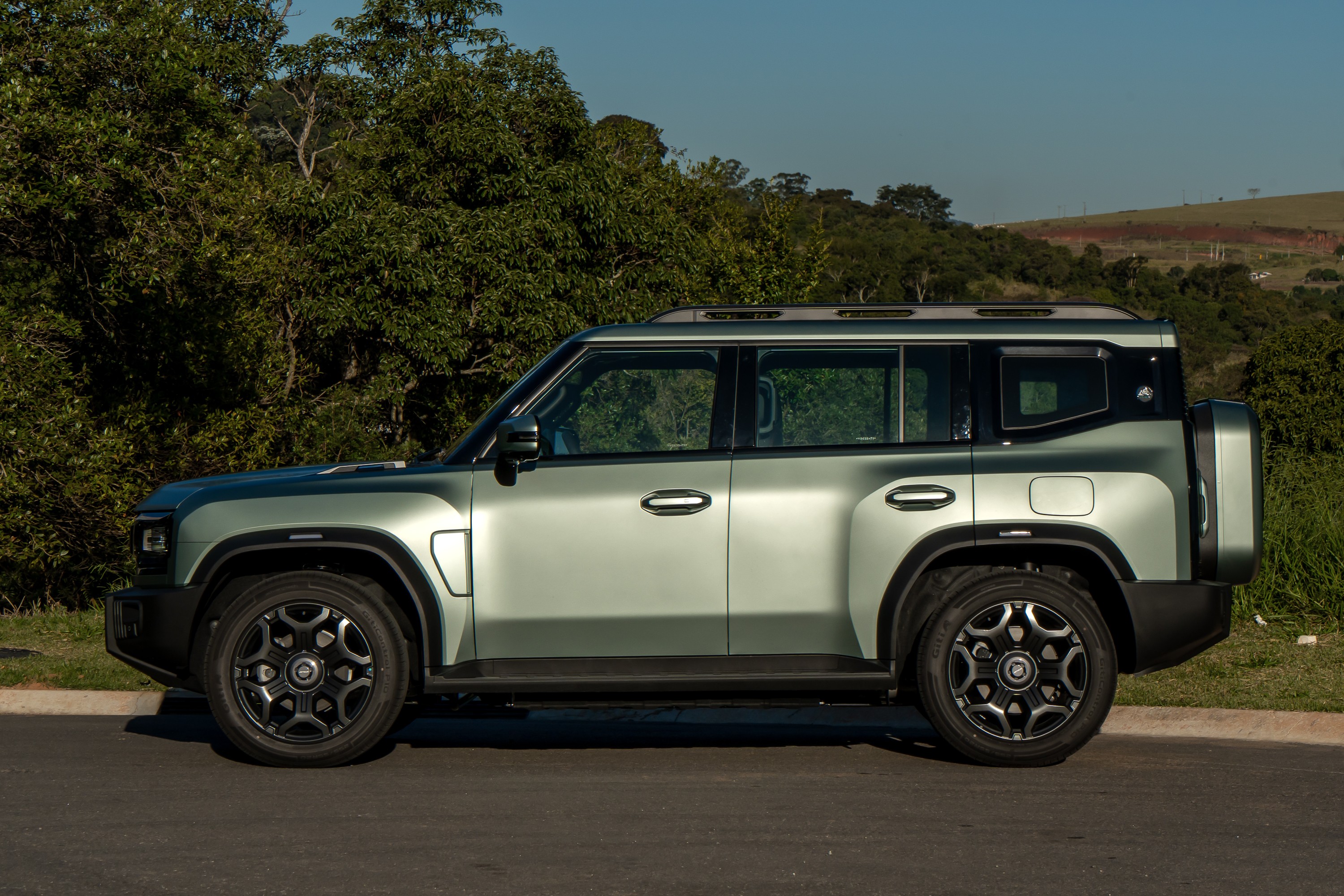 SUV verde-claro com teto preto e rack de teto, estacionado de lado em uma estrada asfaltada, com árvores e vegetação ao fundo sob um céu azul