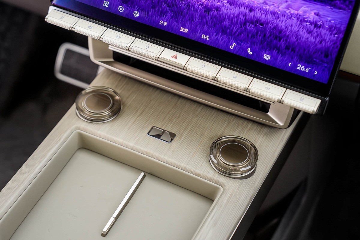 Painel de controle de carro com tela sensível ao toque exibindo um fundo roxo, botões físicos prateados e dois botões giratórios transparentes. Uma caneta stylus prateada repousa em um compartimento abaixo