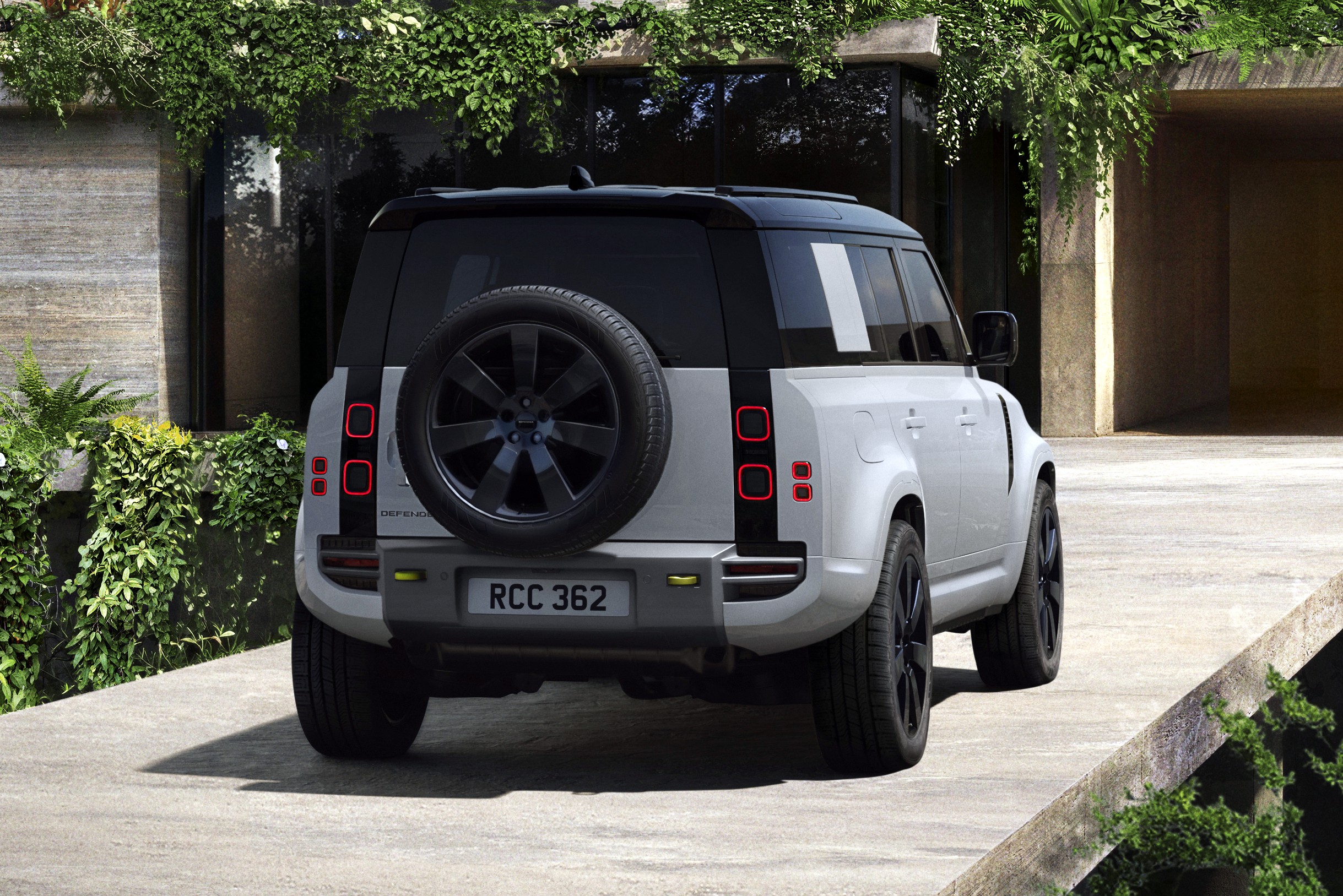 Traseira de um Land Rover Defender branco com teto preto, pneu sobressalente na porta traseira e lanternas quadradas vermelhas, estacionado em uma rampa de concreto com vegetação exuberante em um edifício moderno ao fundo