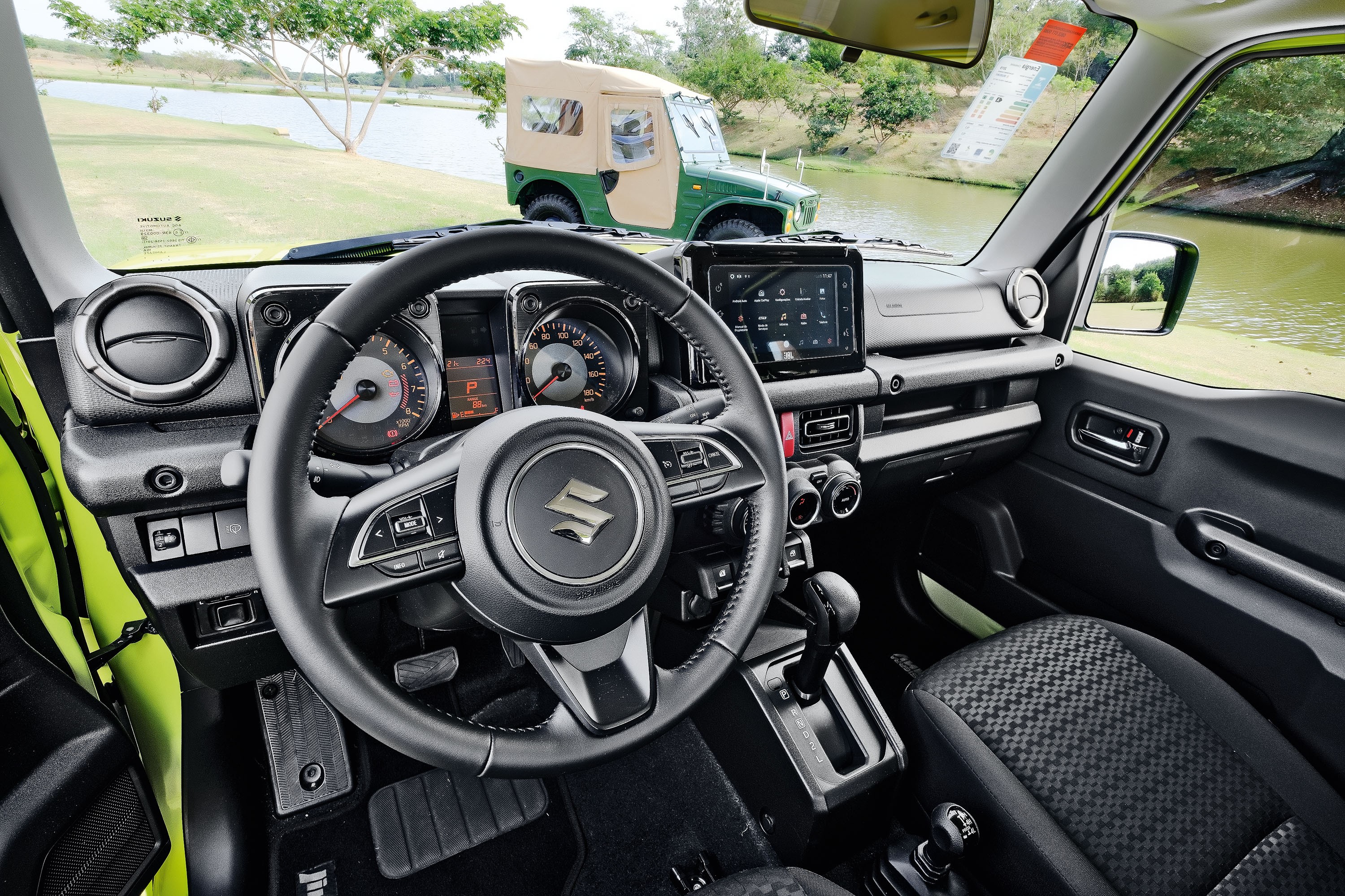 Interior de um carro Suzuki Jimny preto, com volante e painel de instrumentos visíveis. Ao fundo, um jipe verde e bege está parado próximo a um lago, com árvores e gramado ao redor