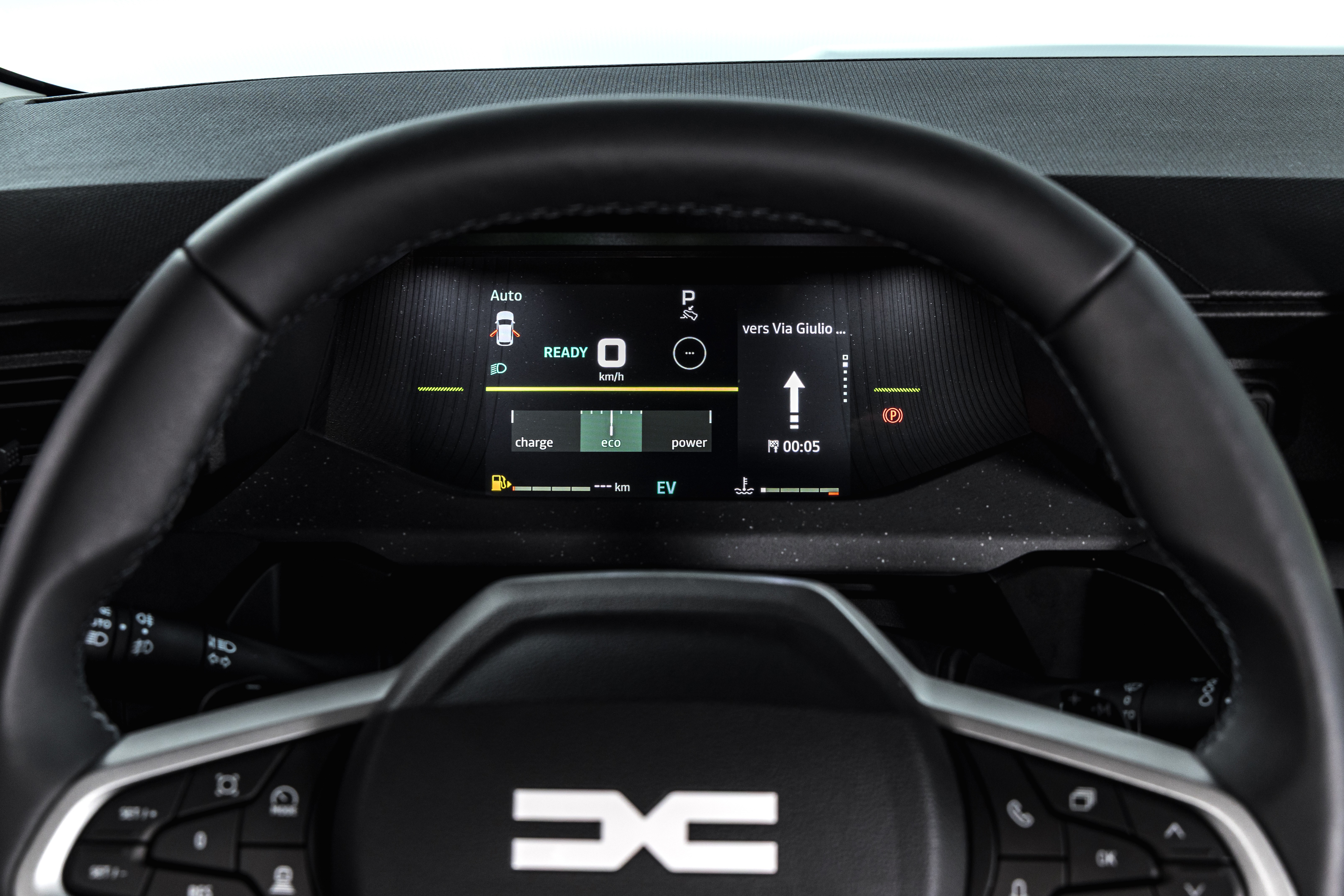 Painel digital de um carro elétrico Dacia, com volante preto em primeiro plano. O painel exibe READY, 0 km/h, indicadores de carga e modo eco, e navegação para Via Giulio com seta para cima e tempo estimado de 00:05. O logo Dacia está no centro do volante